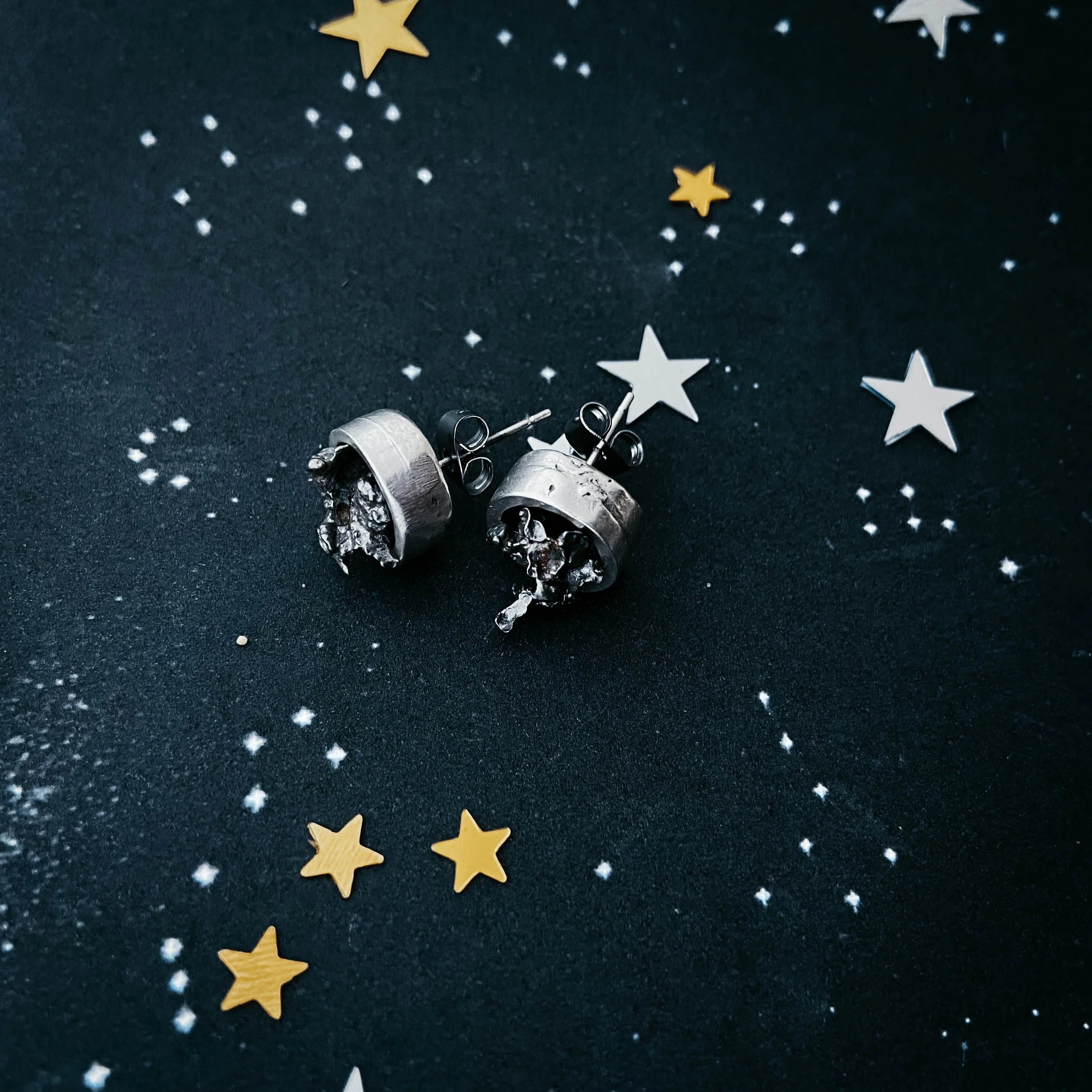 Silver stud earrings with organic textured design on black starry background with gold and white star confetti