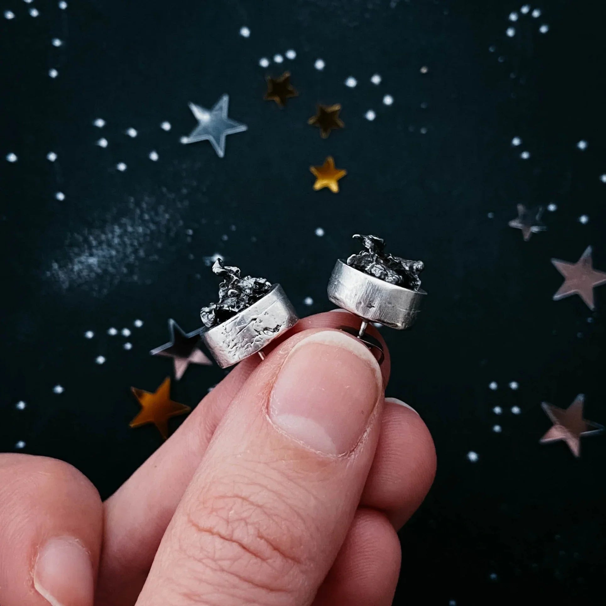 Hand holding artist-designed silver earrings with cosmic motif, star confetti background, Qu!rk Boutique