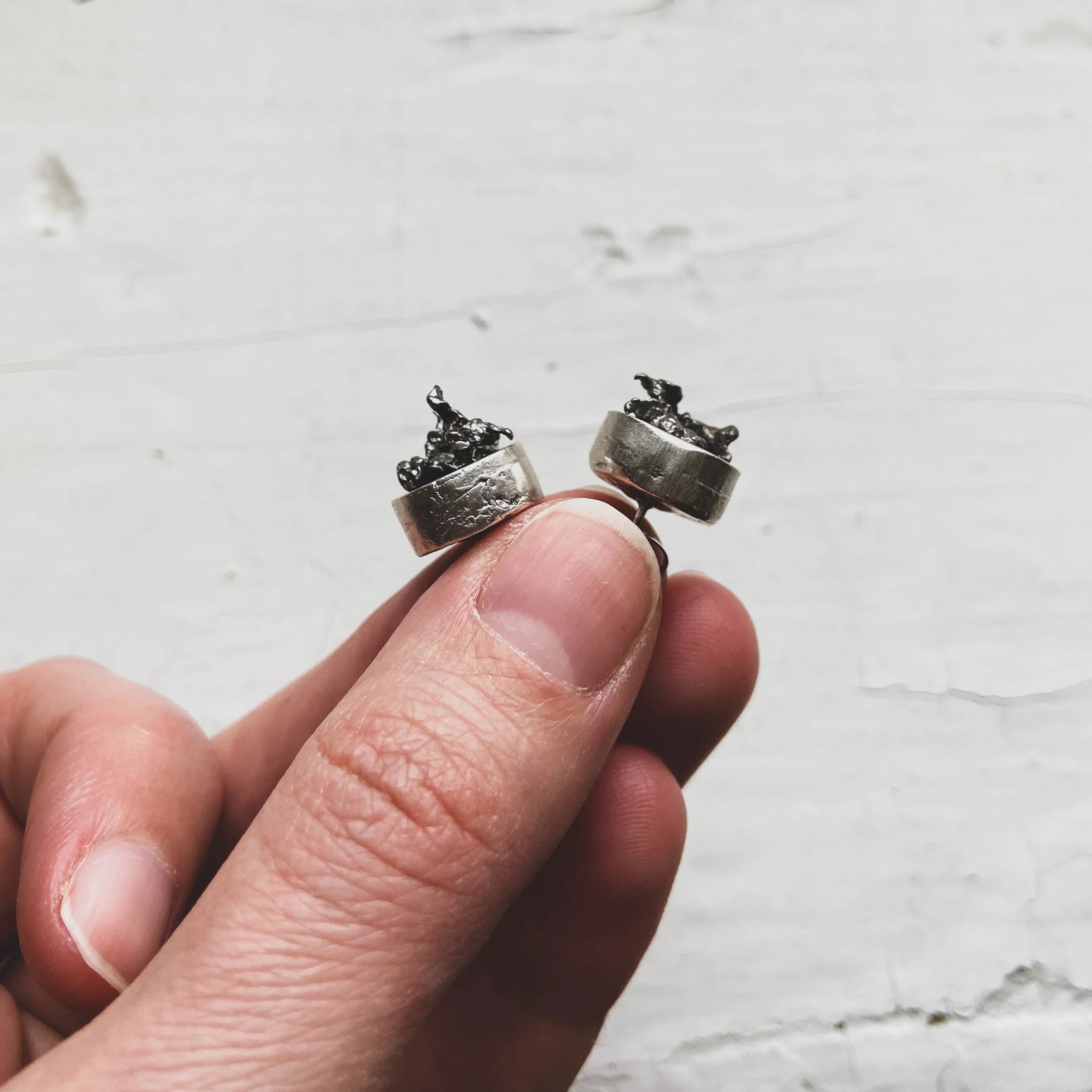 Hand holding a pair of silver stud earrings with dark, textured, organic-shaped tops