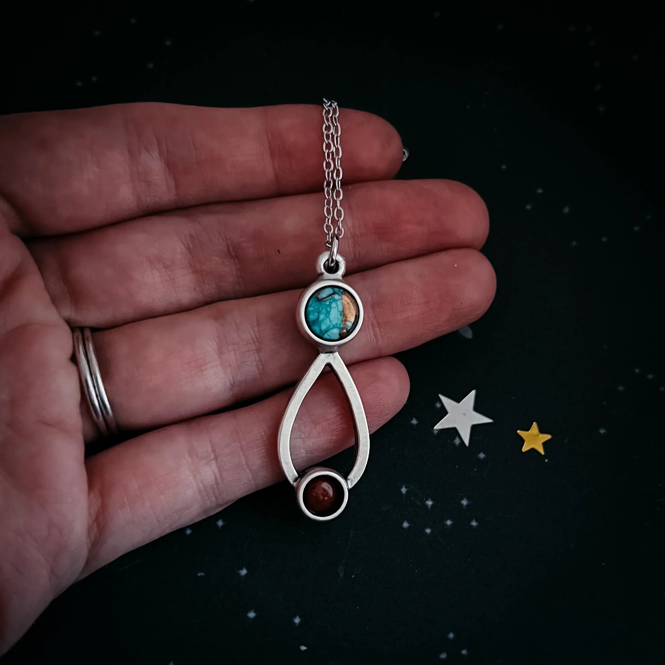 Hand holding silver necklace pendant with turquoise and brown stones on black background with star confetti