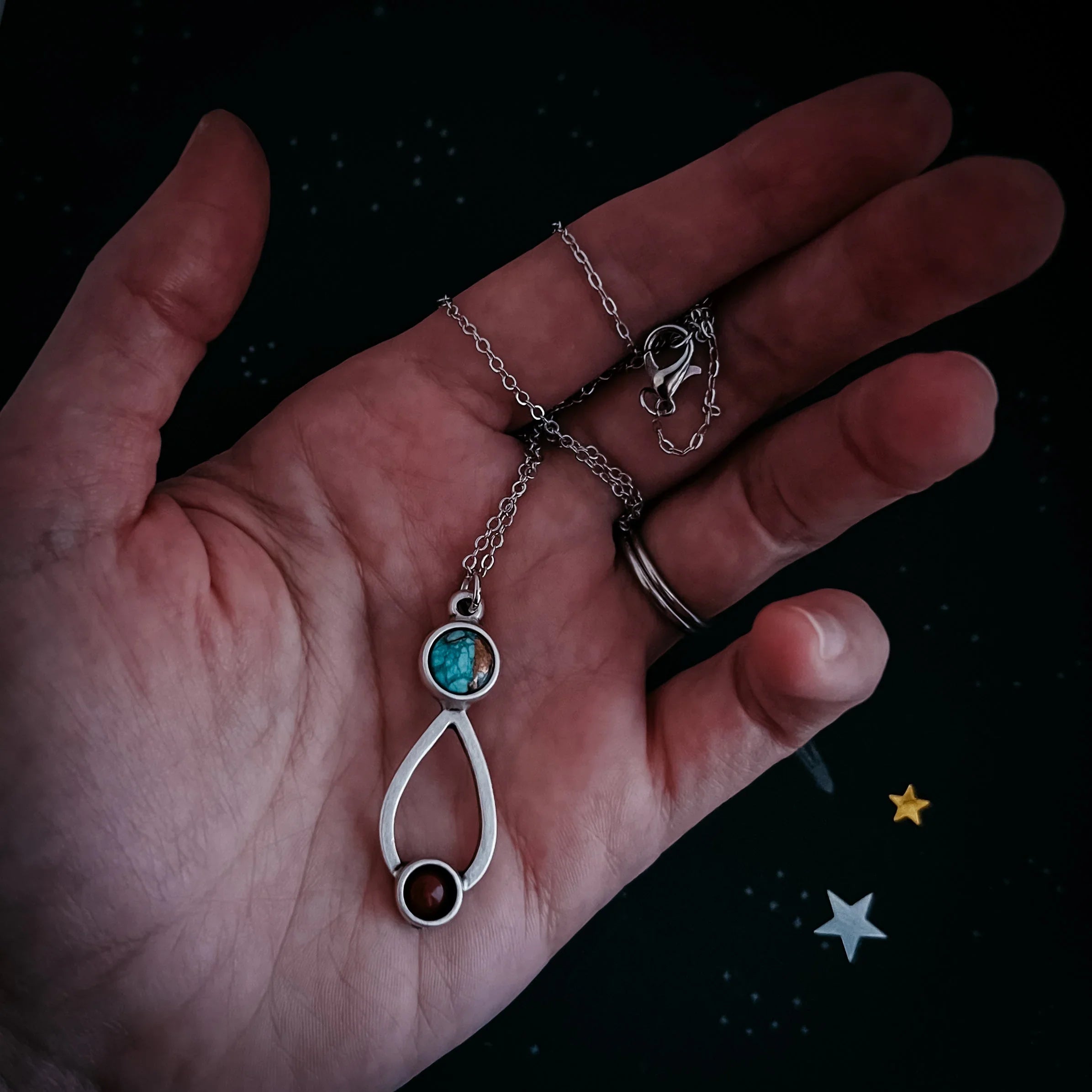 Hand holding silver necklace with turquoise and brown stone pendant on star-patterned black fabric
