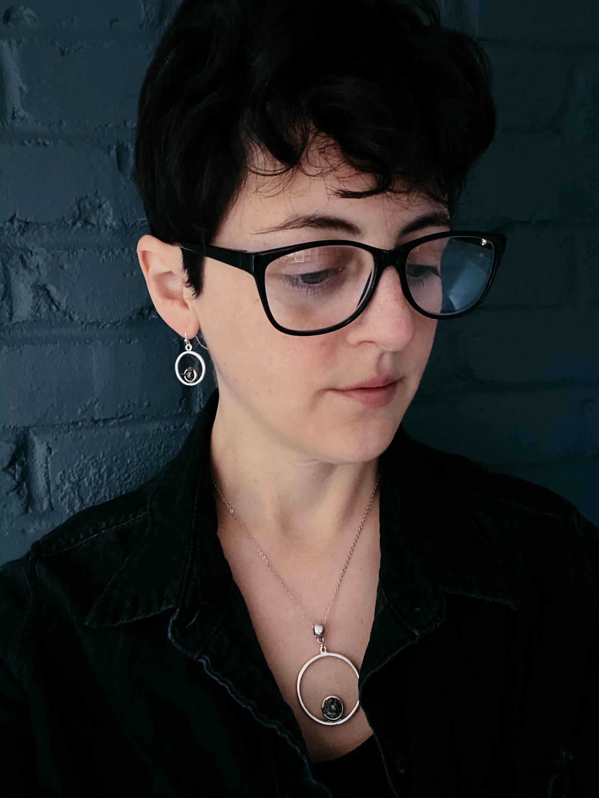 Woman with short dark hair and glasses wearing silver circle earrings and matching necklace against dark brick wall