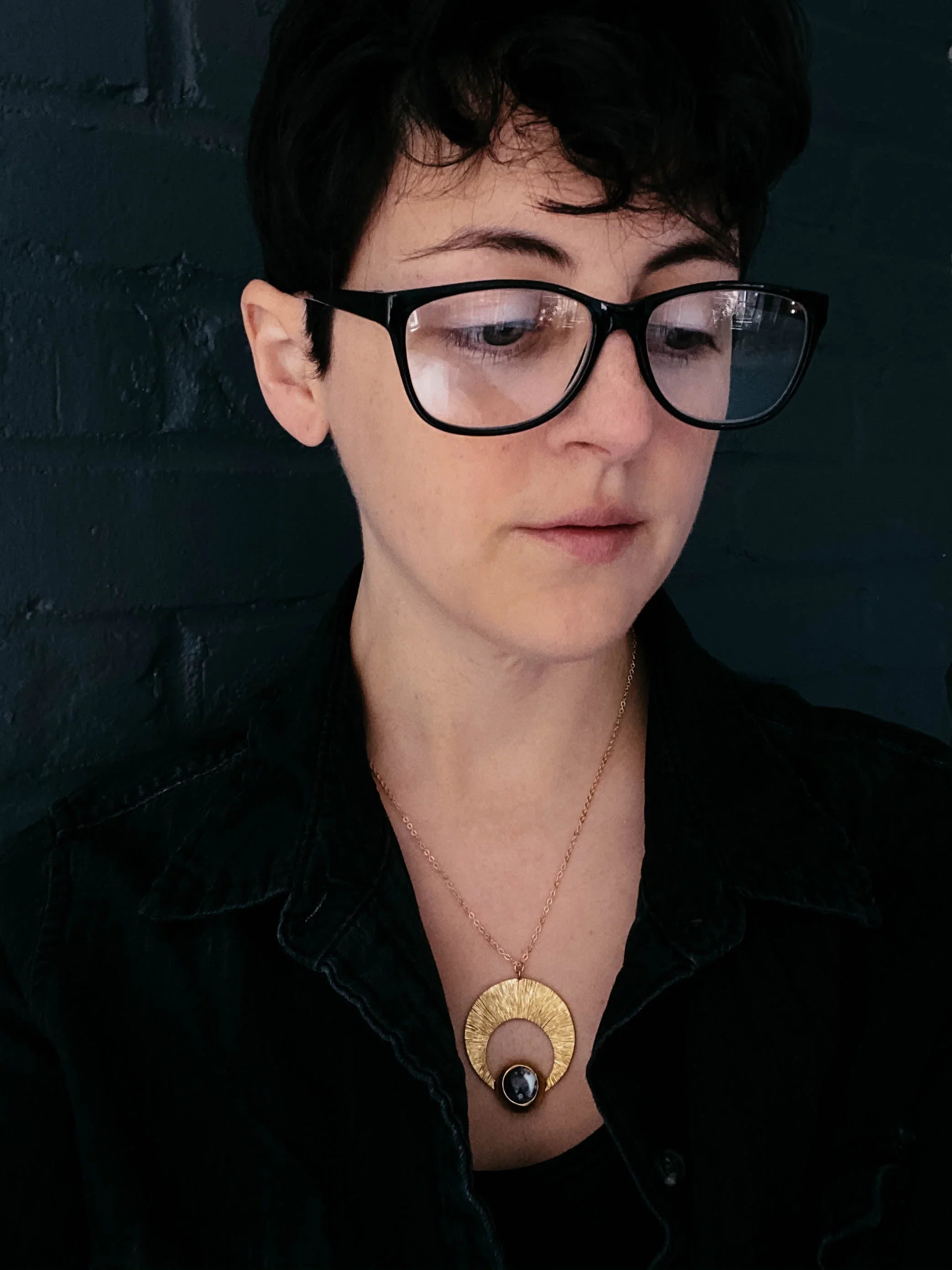 Person wearing glasses and a bold gold crescent pendant necklace, artist-designed jewelry, Qu!rk Boutique