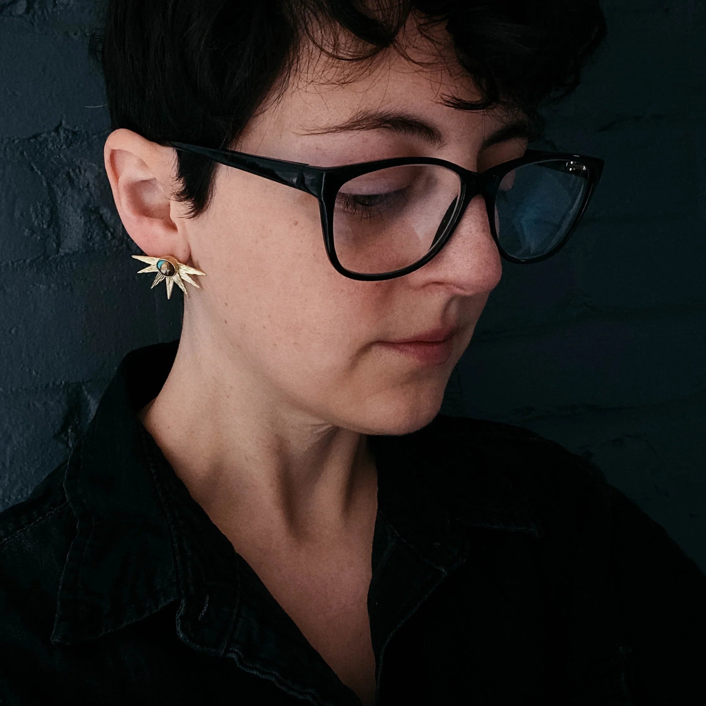 Woman with short dark hair wearing black glasses and gold sunburst earrings against dark background