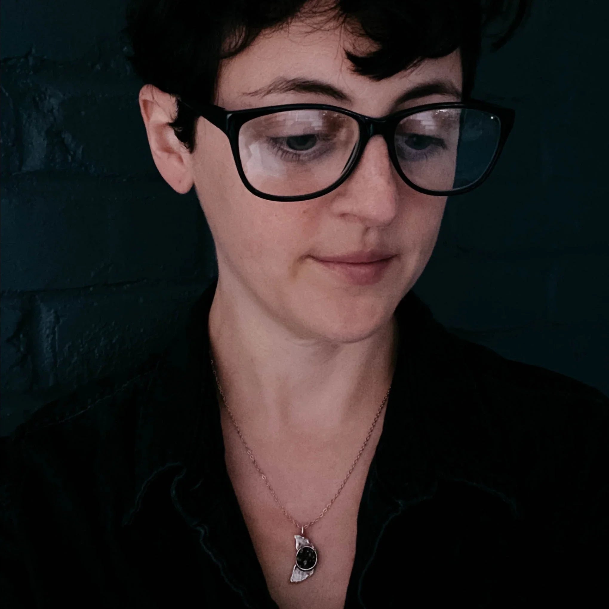 Close-up of a person with short dark hair, black glasses, and a silver necklace with a black pendant against a dark wall