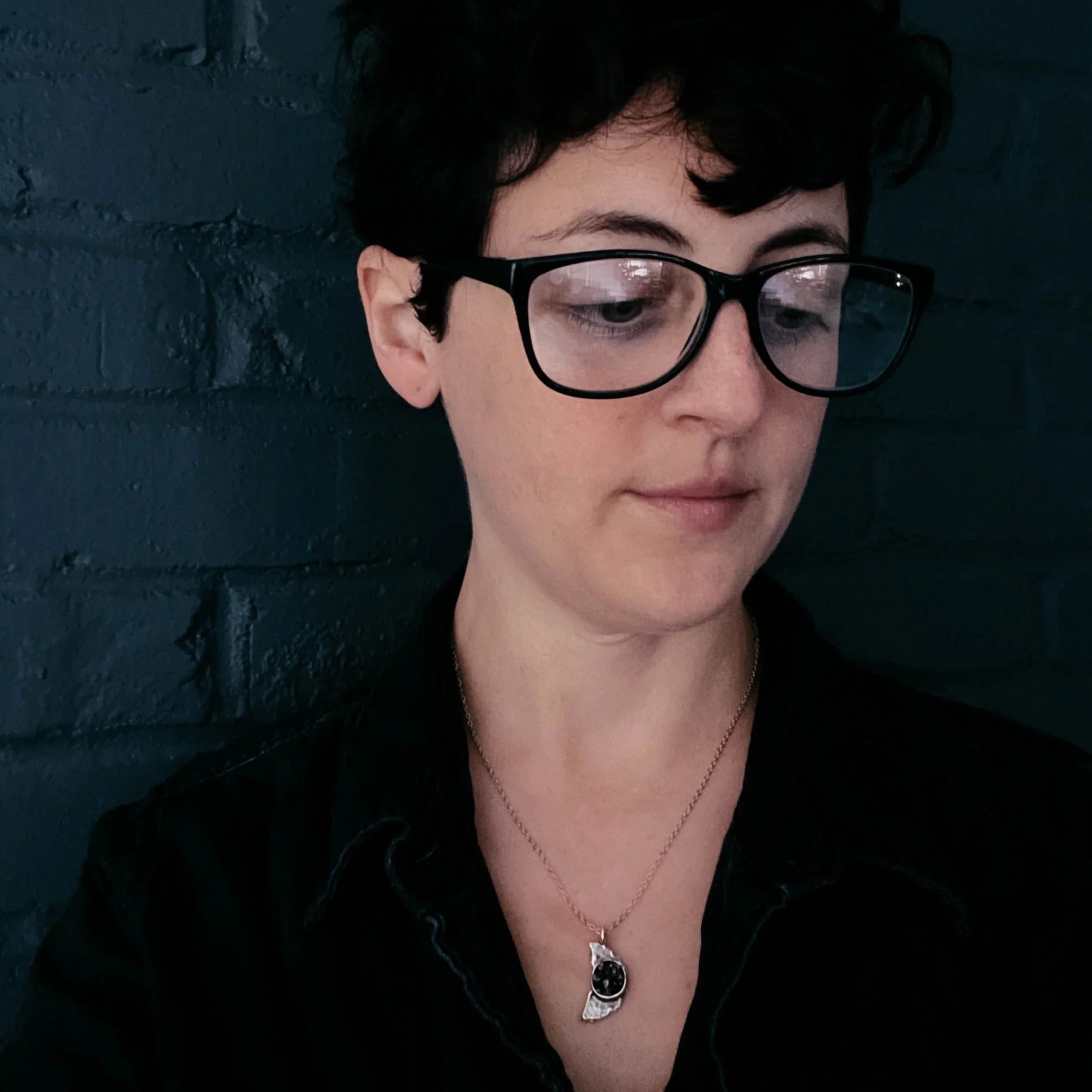 Person with short dark hair and large glasses wearing a moon-shaped pendant necklace against dark brick wall