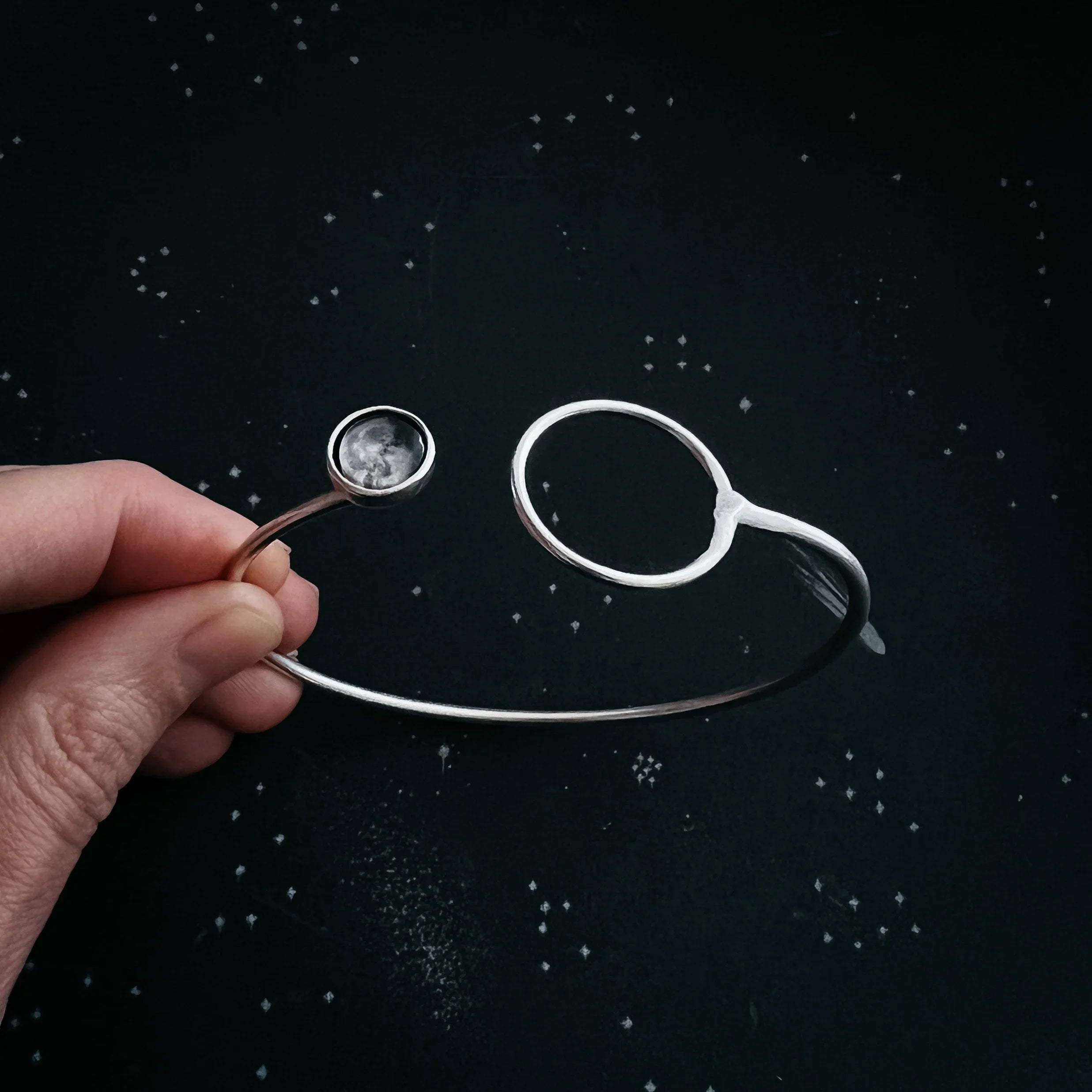 Hand holding silver cosmic-inspired open cuff bracelet with moon detail on starry black background