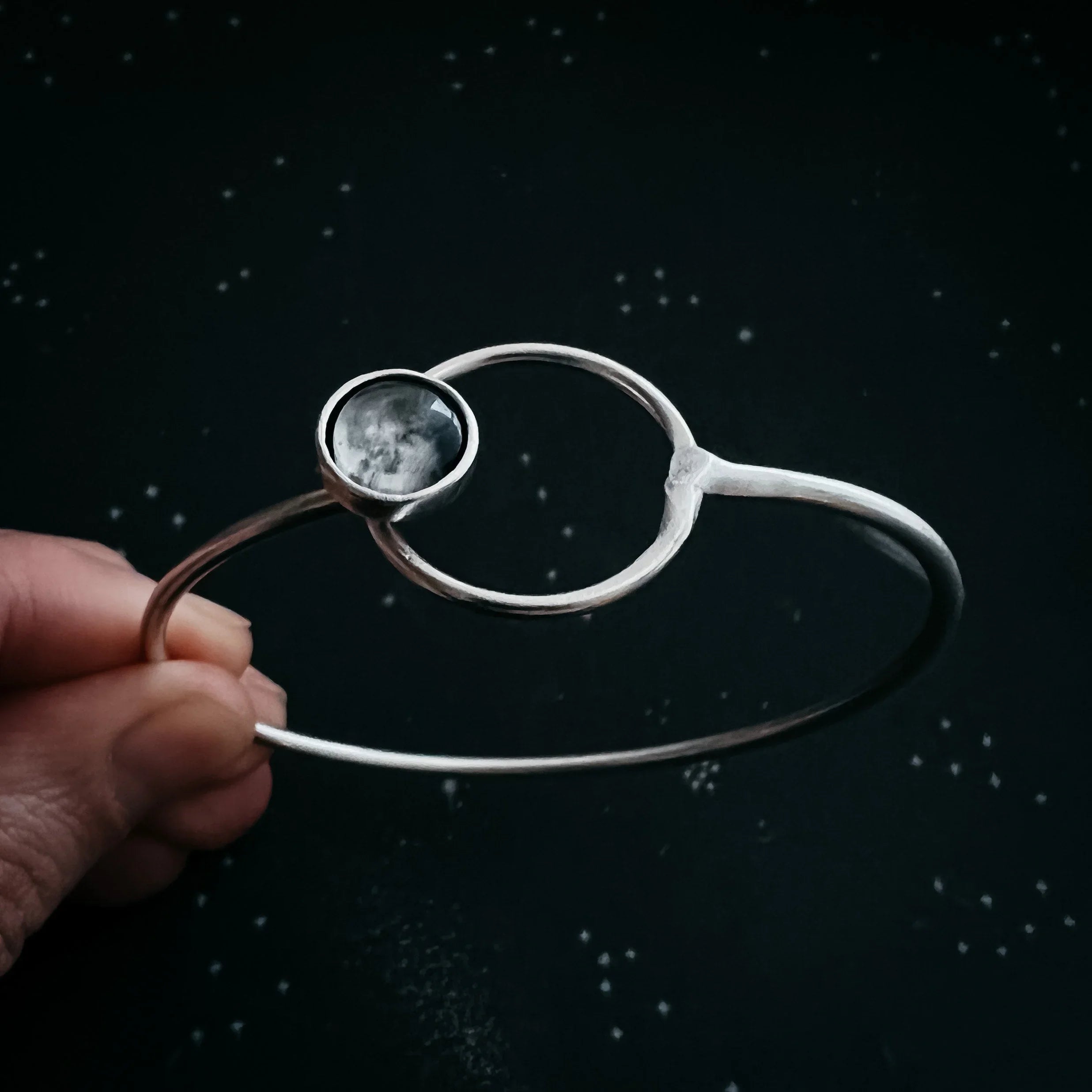 Hand holding a silver moonstone bracelet with cosmic design on a starry black background