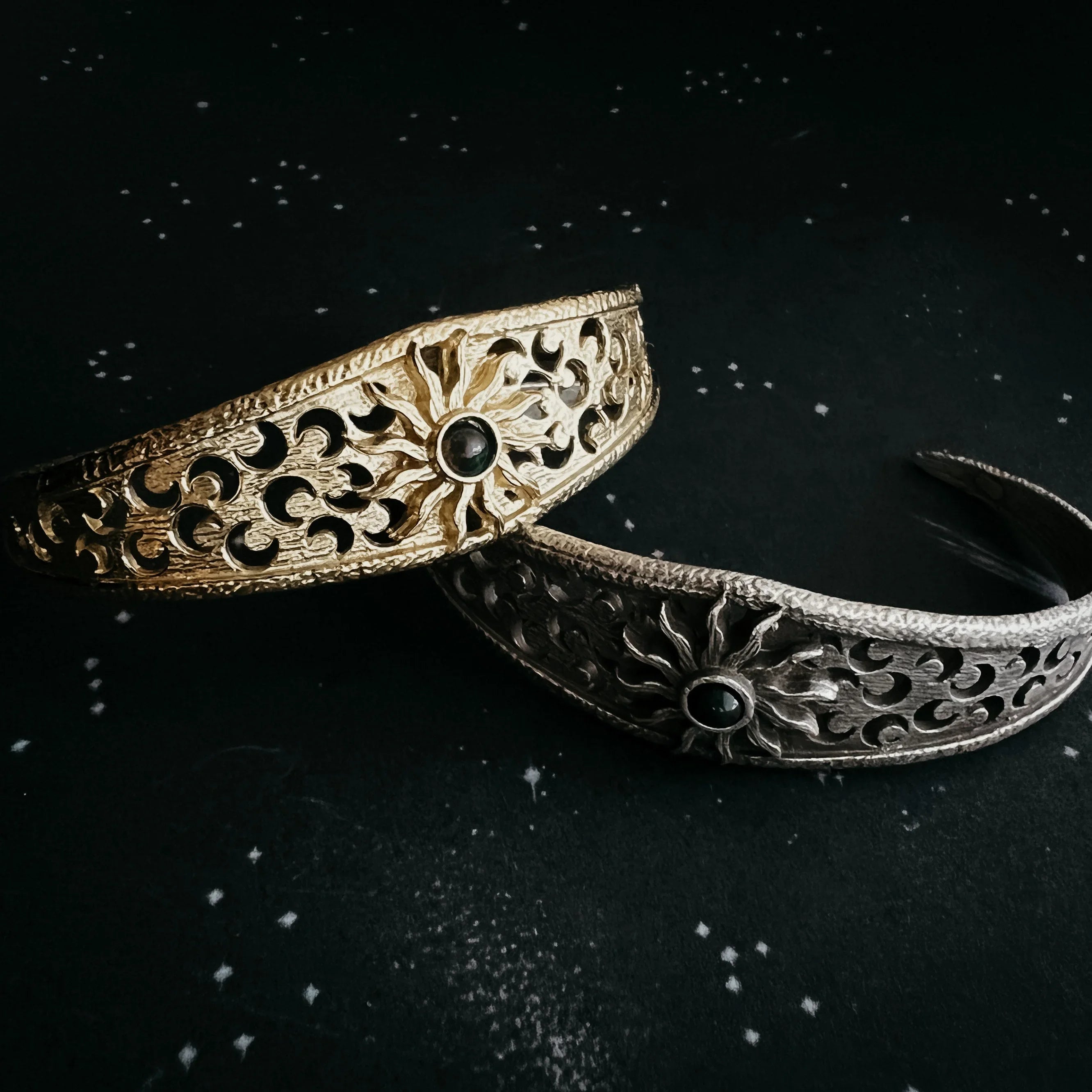 Gold and silver sunburst cuff bracelets with cosmic detailing on a star-speckled black background