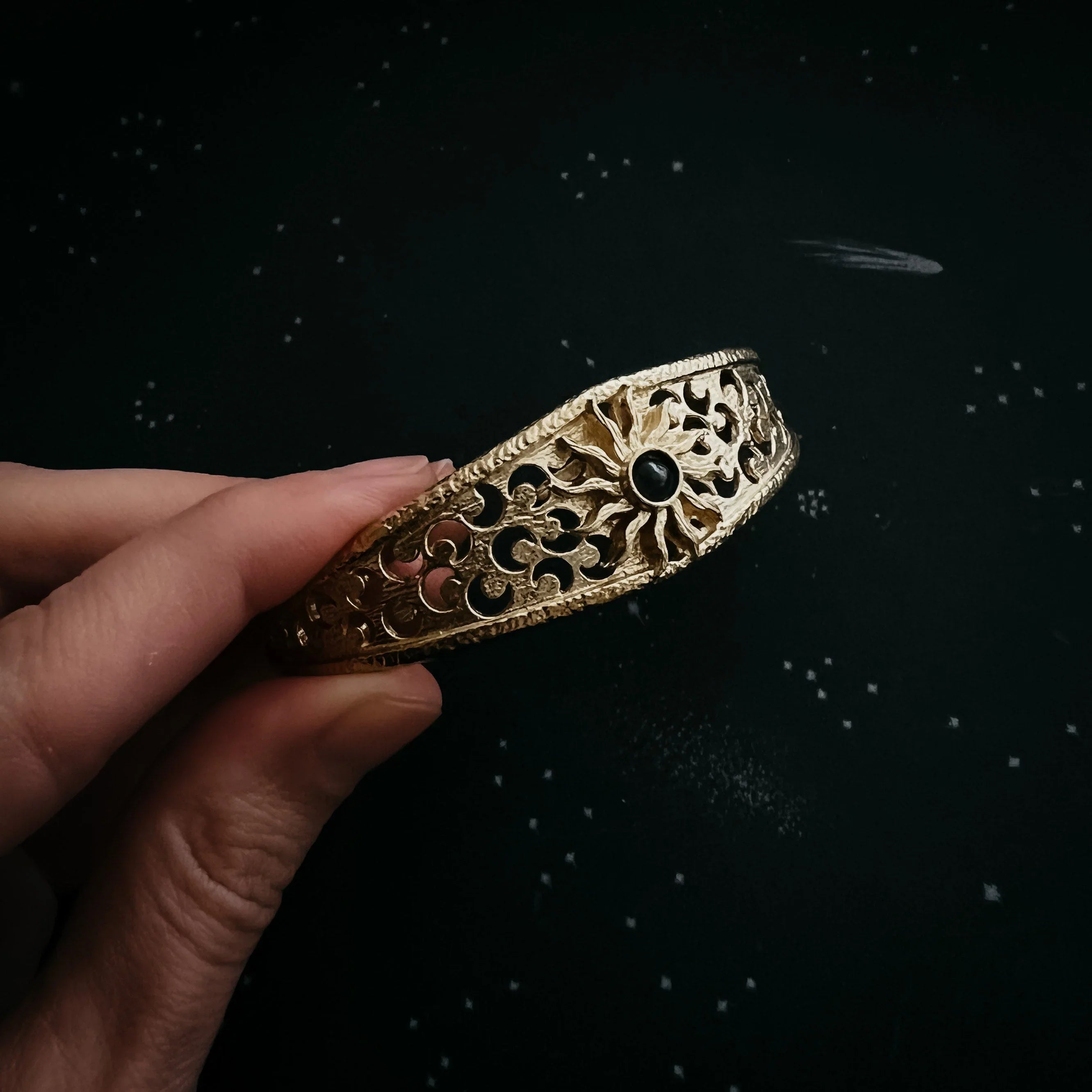 Hand holding ornate gold bracelet with sun design and black stone on dark starry background