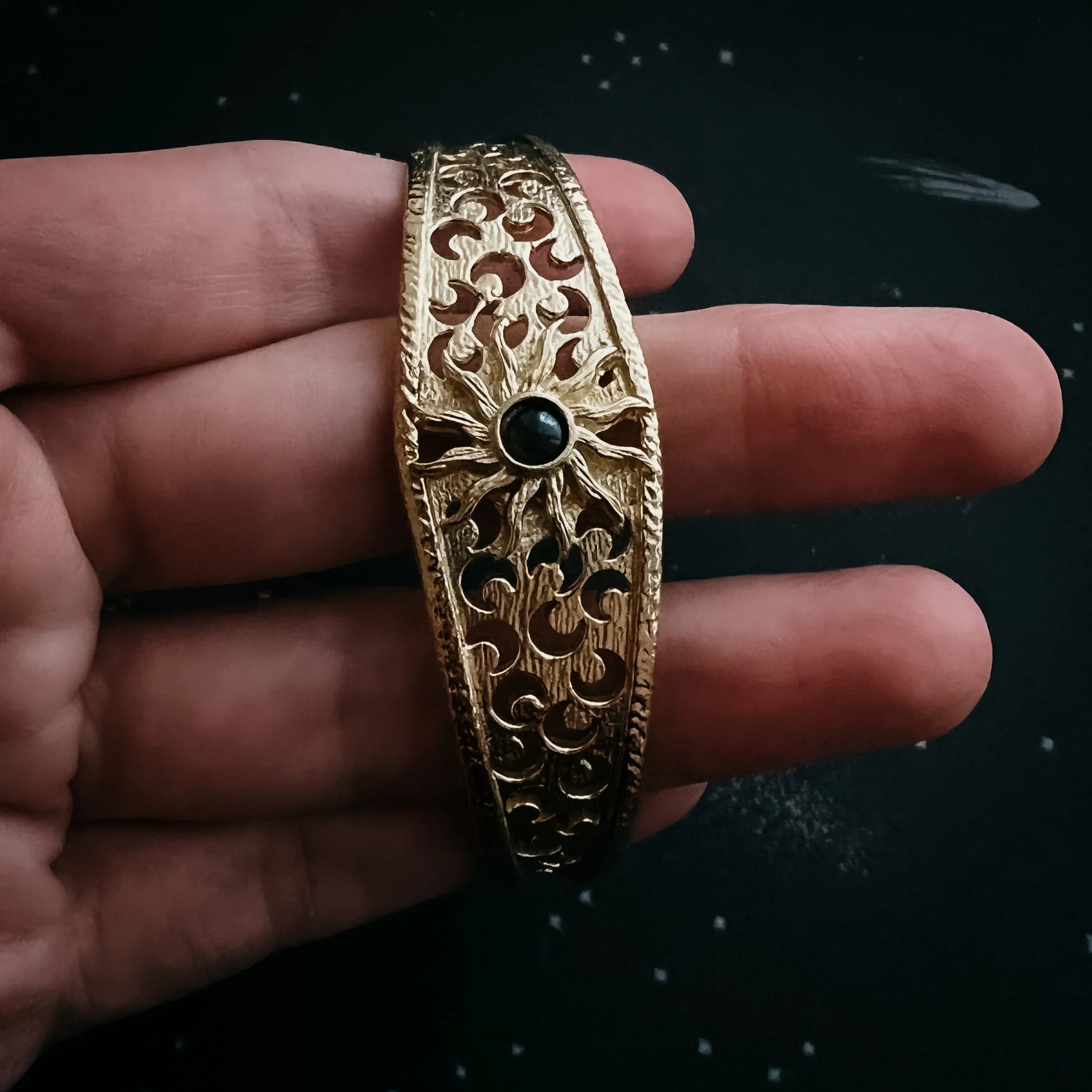Hand holding ornate gold bracelet with sun design and black stone accent on dark background