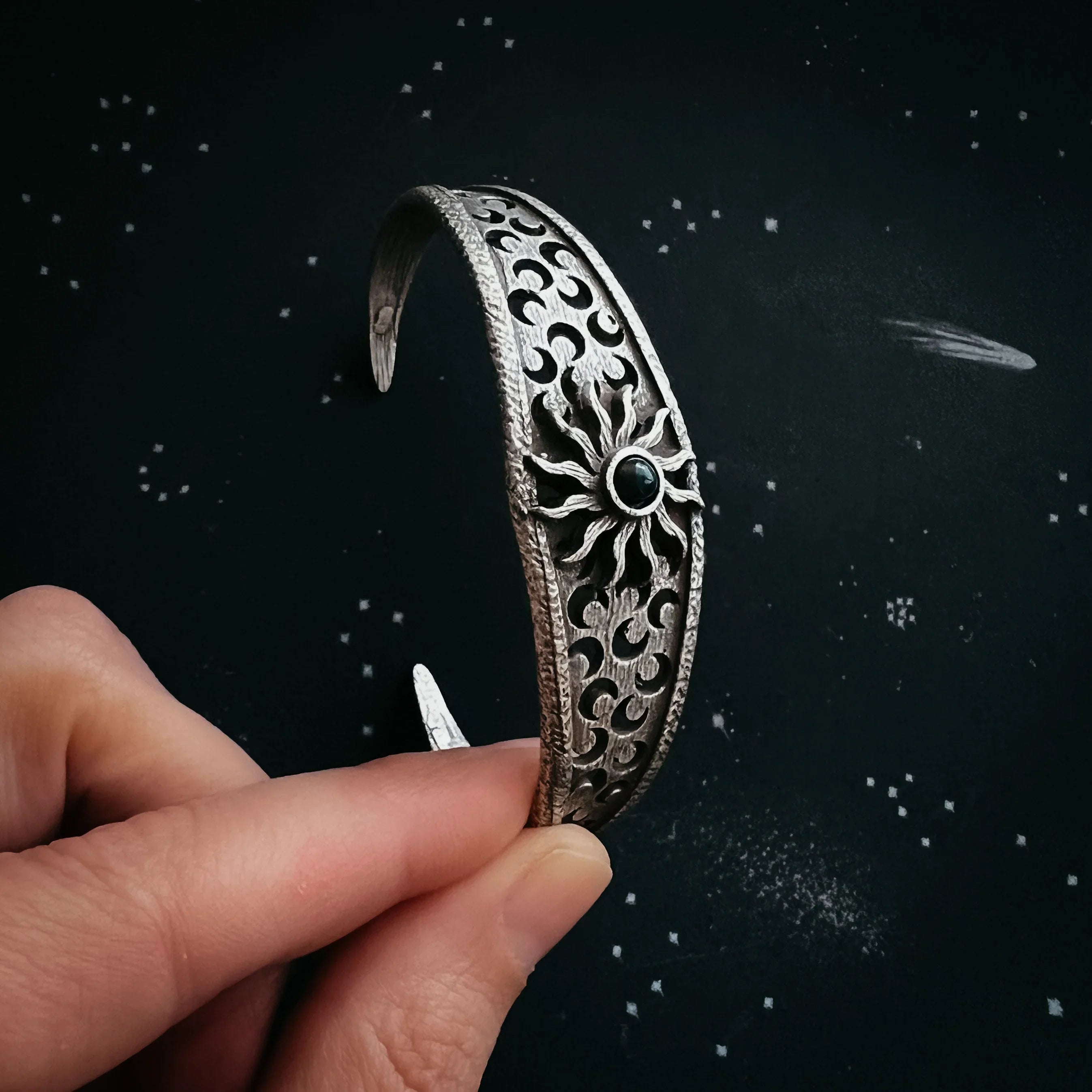 Hand holding a silver cuff bracelet with sun and wave cutout design on a starry black background