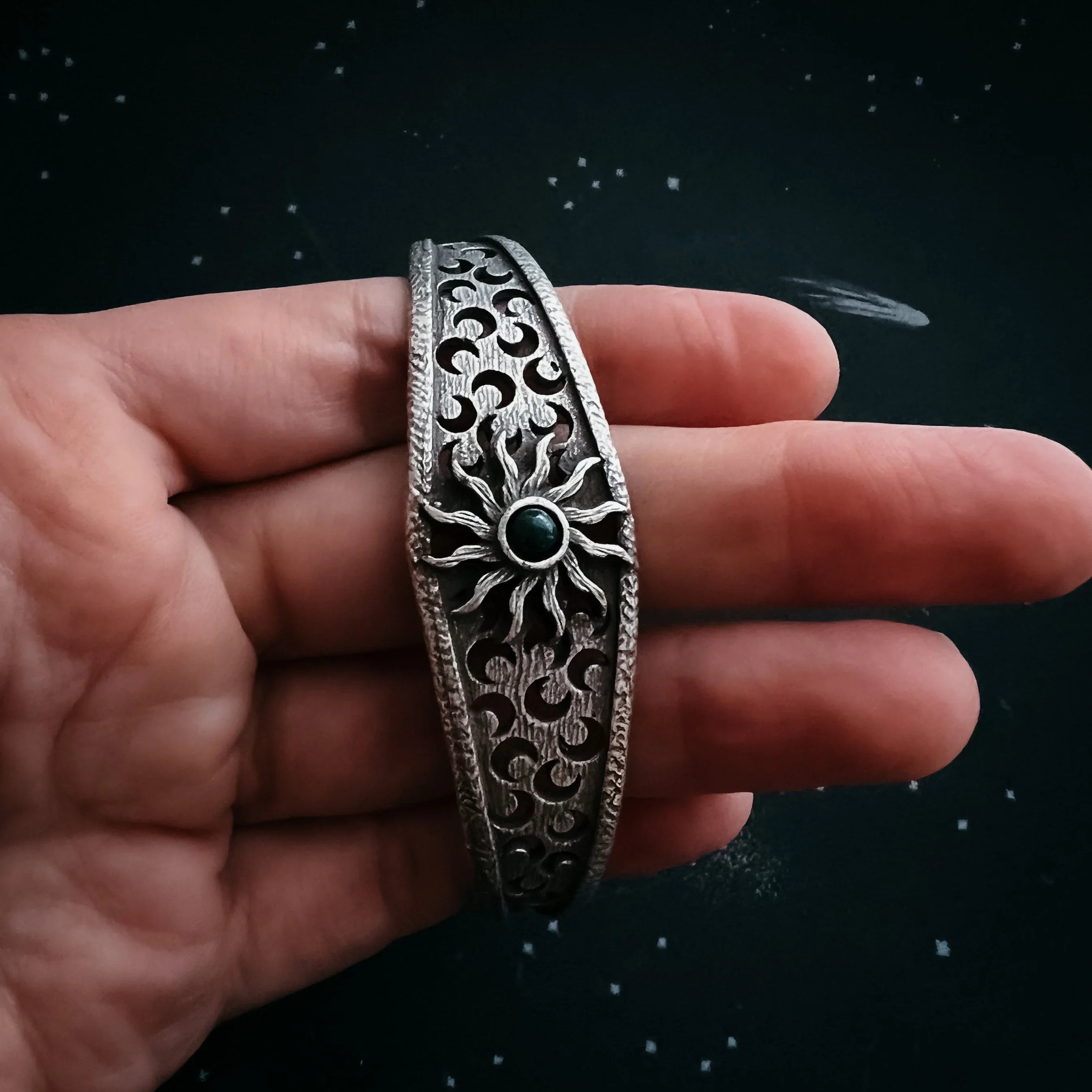 Hand holding an ornate silver bracelet with sun and moon cut-out designs and a green stone center