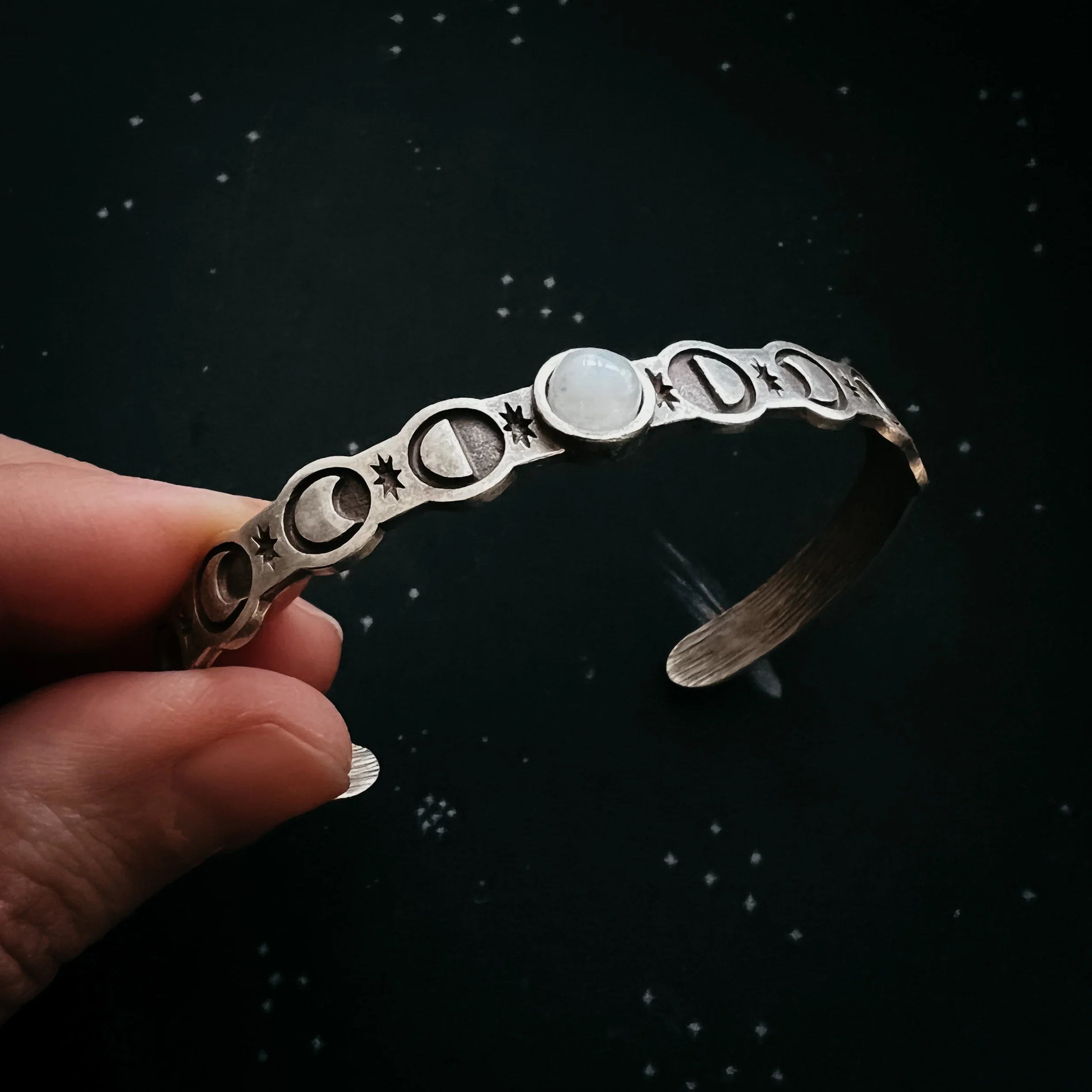 Hand holding a silver cuff bracelet with moon phases and gemstone, cosmic background