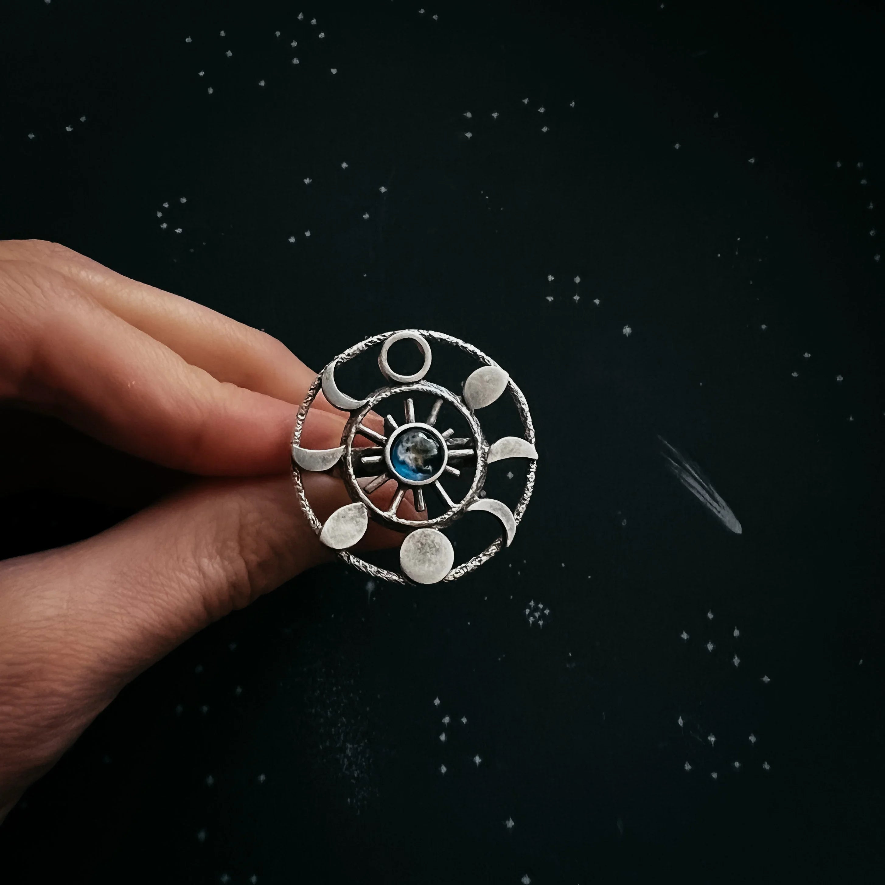 Hand holding silver ring with moon phases and earth stone on starry black background