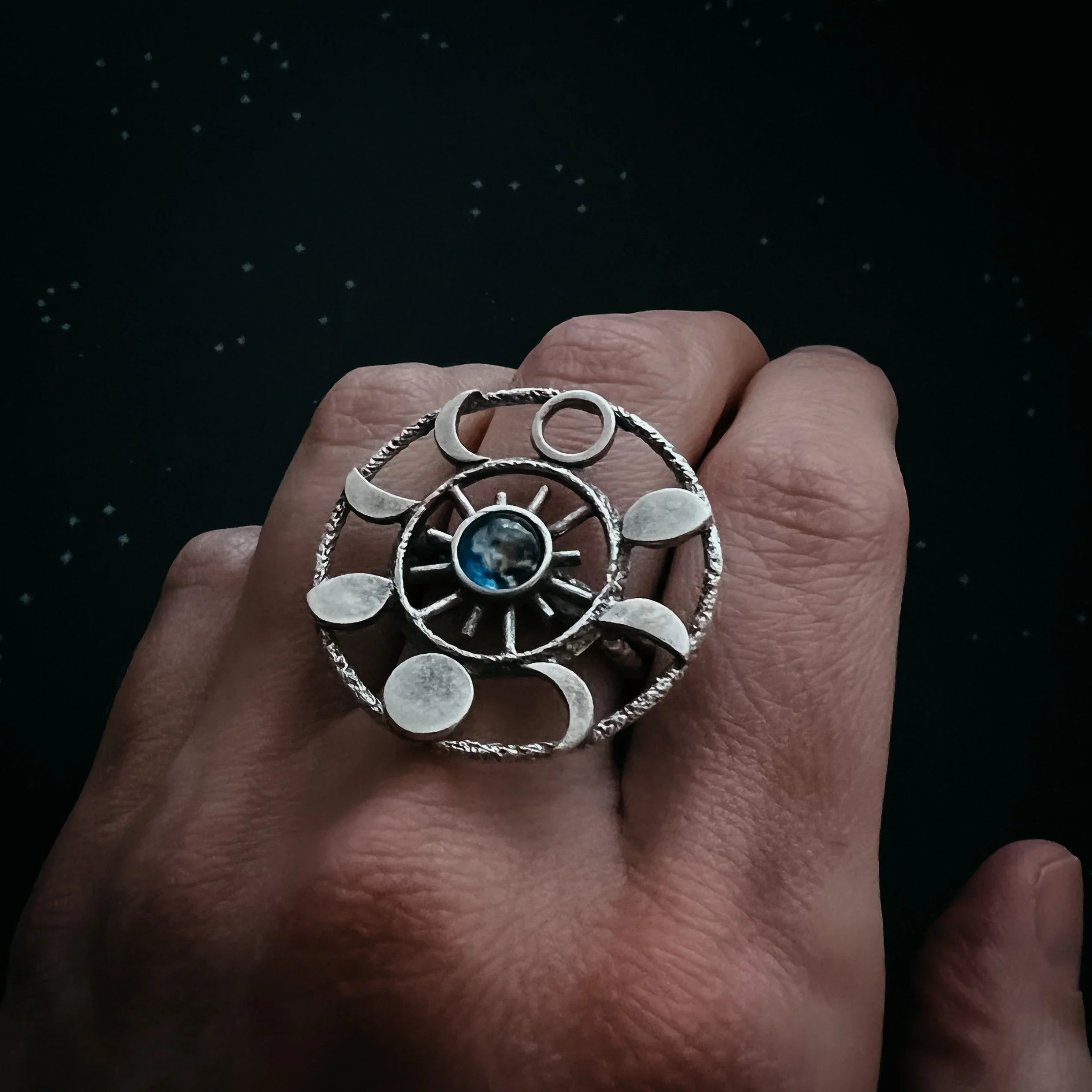 Hand wearing a silver celestial ring with moon phases and a blue stone in the center on a dark starry background