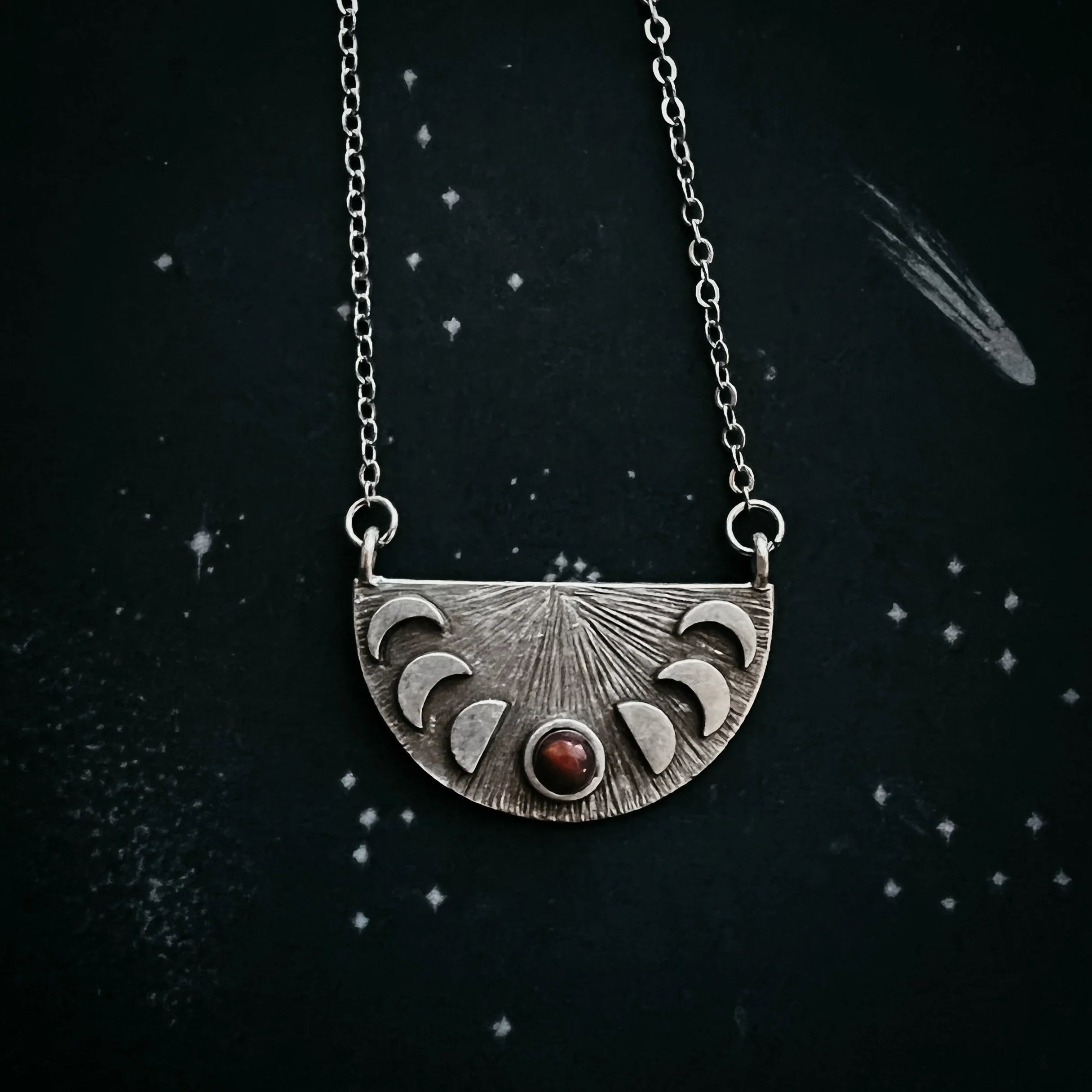 Silver crescent moon phase necklace with red stone on black starry background