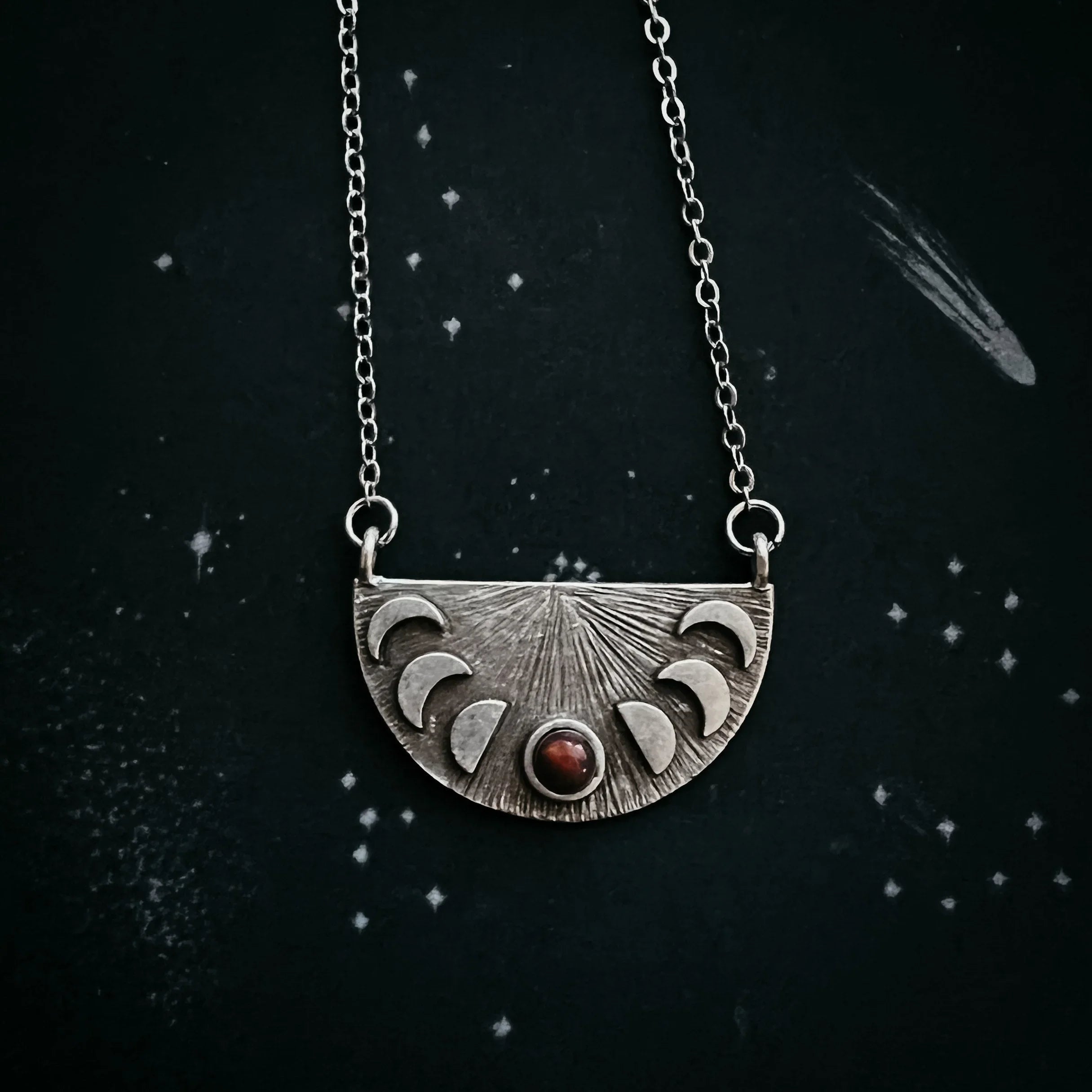Silver necklace pendant with moon phases design and red gemstone on starry black background