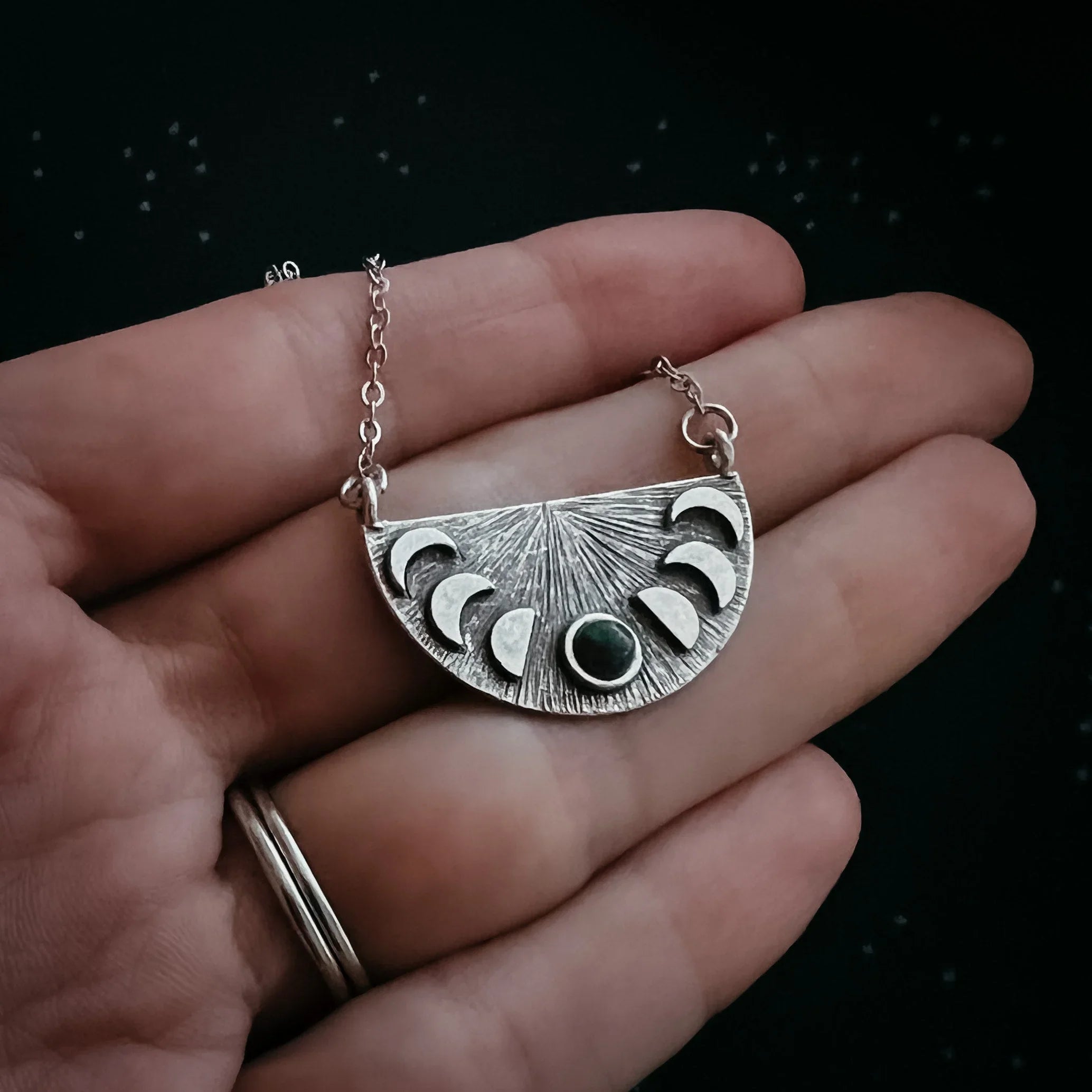 Hand holding a silver necklace pendant with moon phases design and central dark gemstone