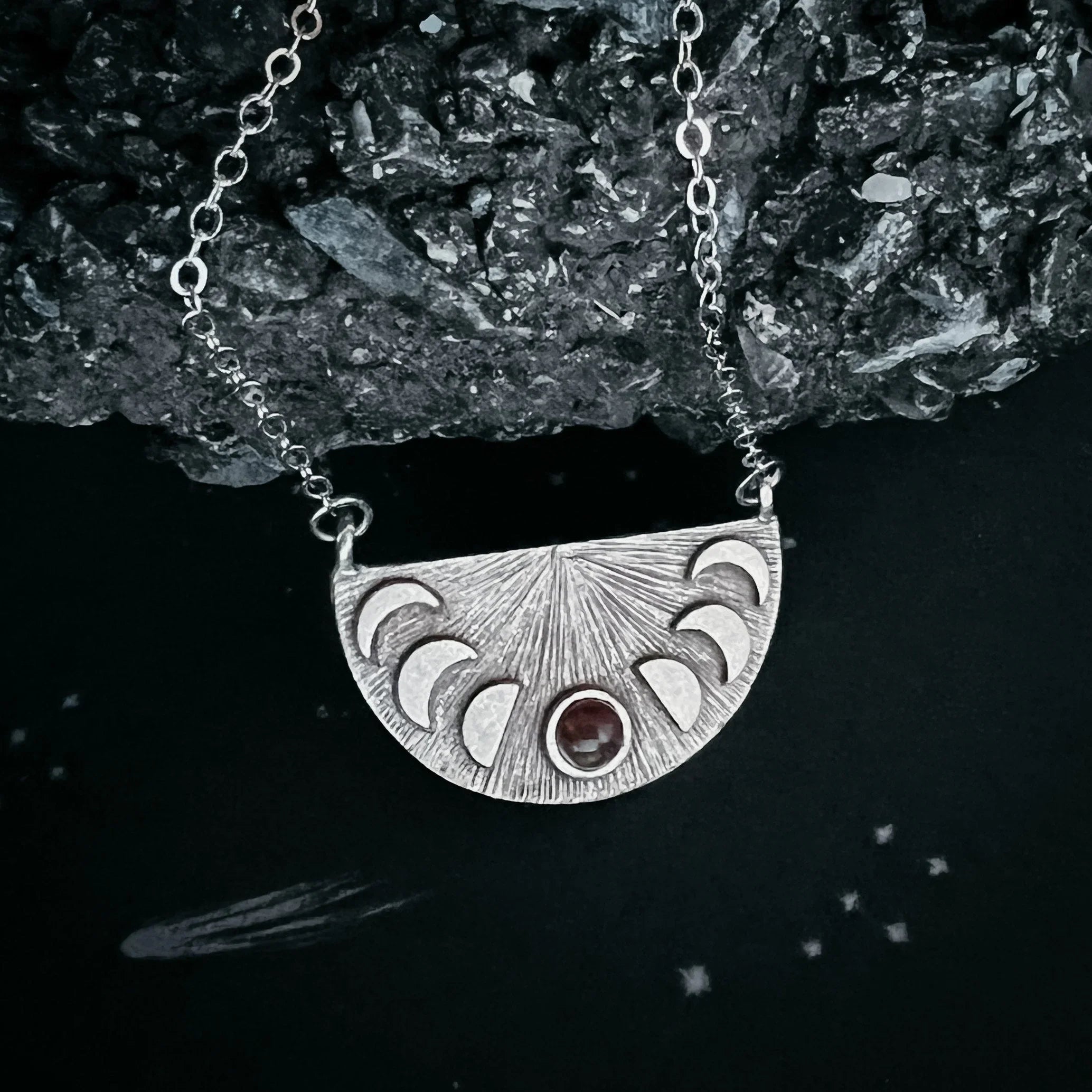 Silver crescent moon phase necklace pendant with textured rays and central red gemstone on chain