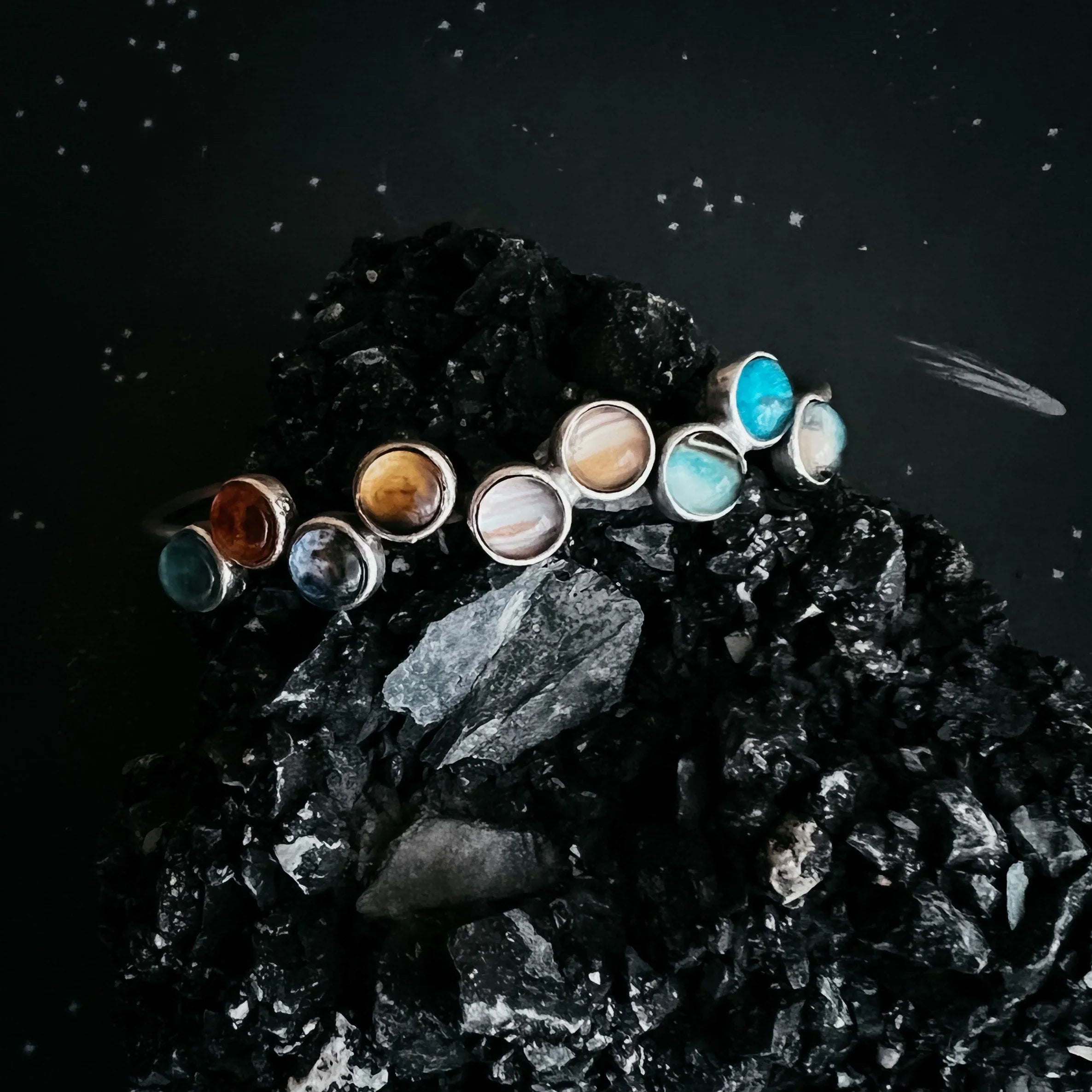 Silver rings with various colorful stones displayed on black textured rocks against a dark background