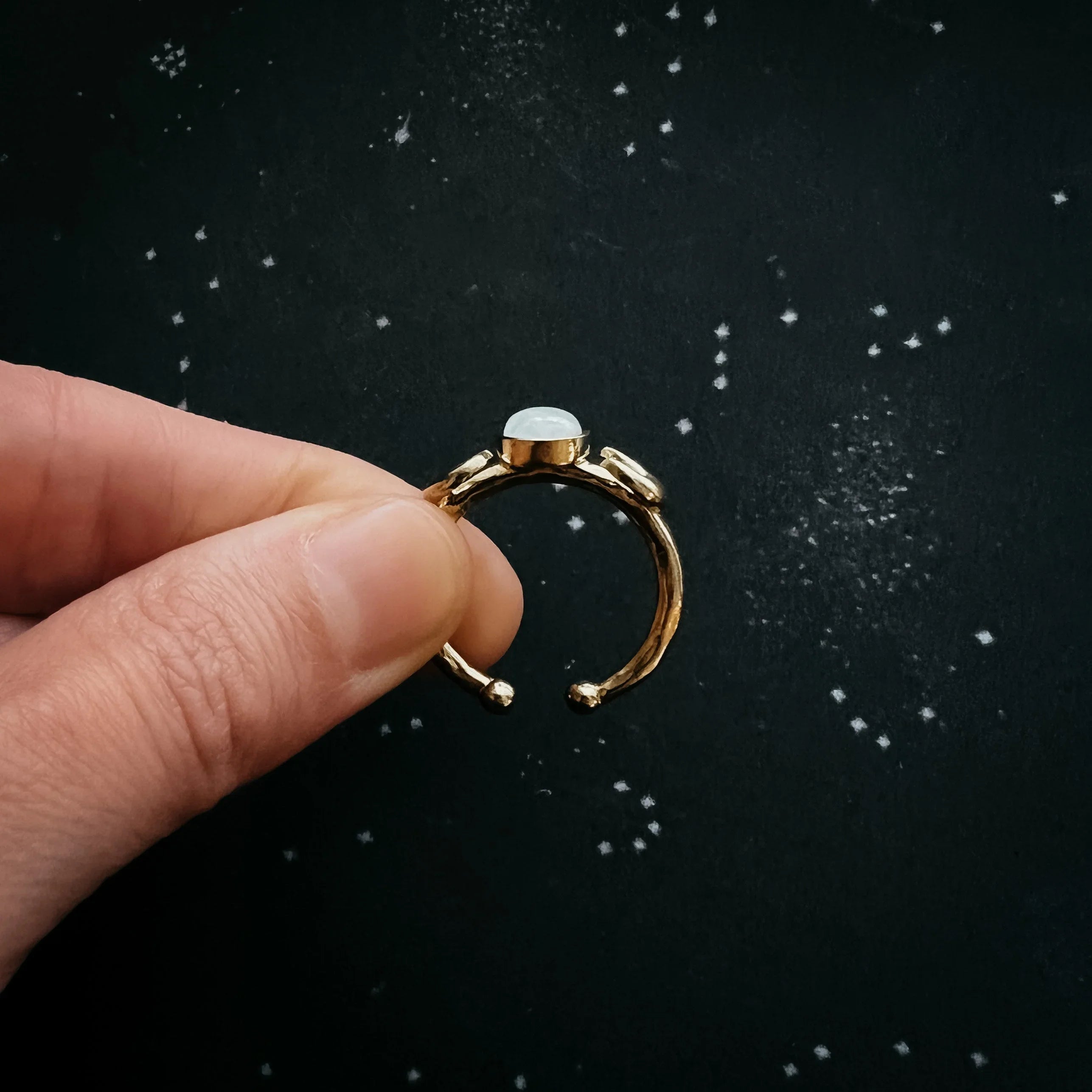 Hand holding a gold open ring with a white stone on a cosmic star-patterned background