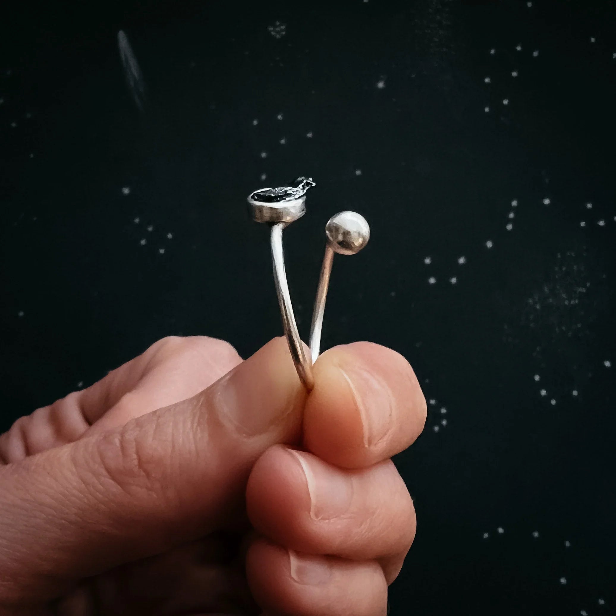 Hand holding a minimalist silver ring with one round and one flat textured end against a dark starry background