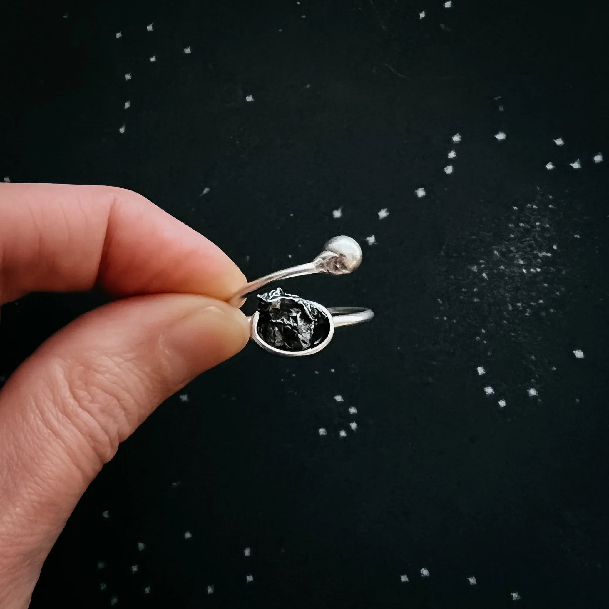 Hand holding a silver open ring with a rough black stone and a small silver ball on a dark background