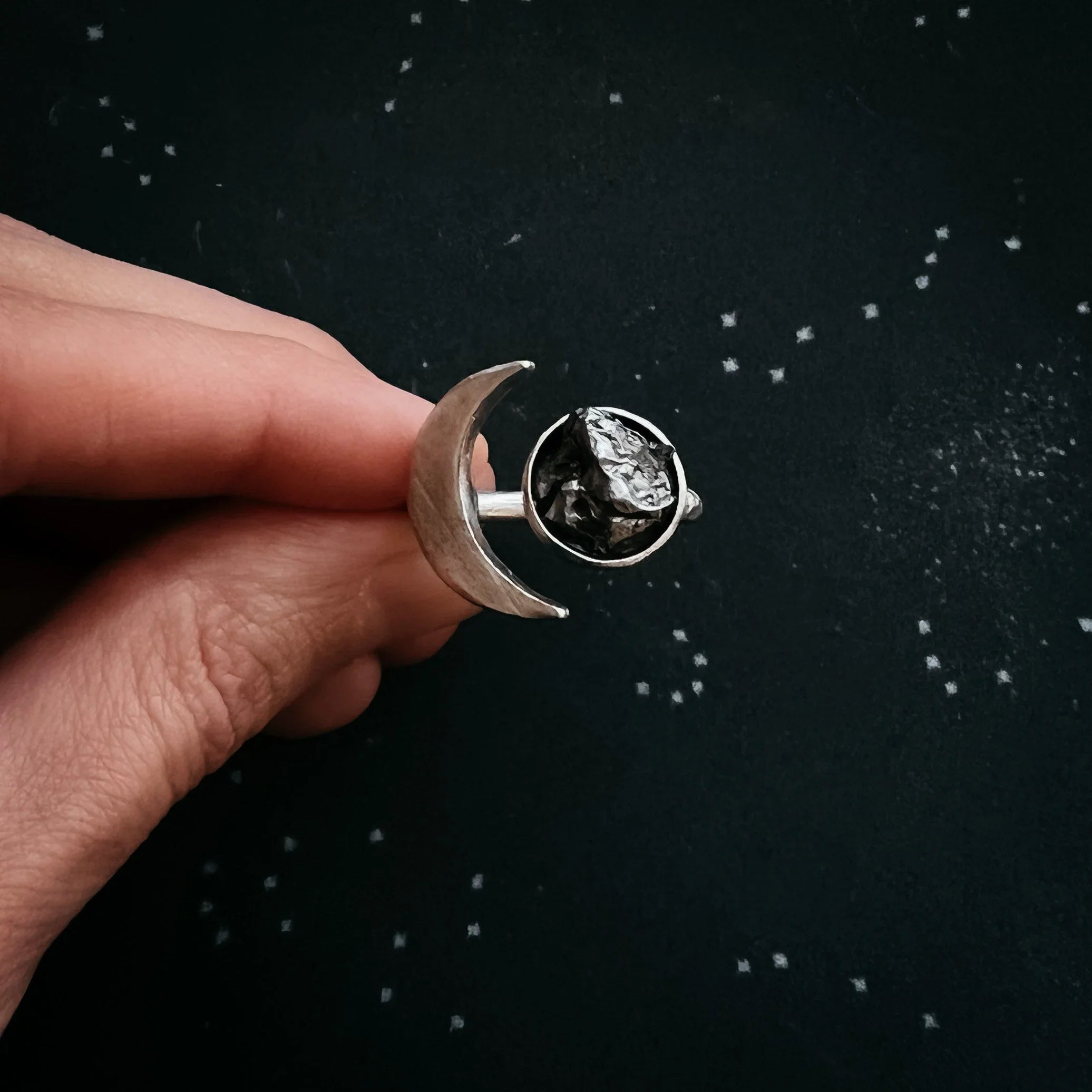 Hand holding silver crescent moon ring with rough black stone on starry black surface