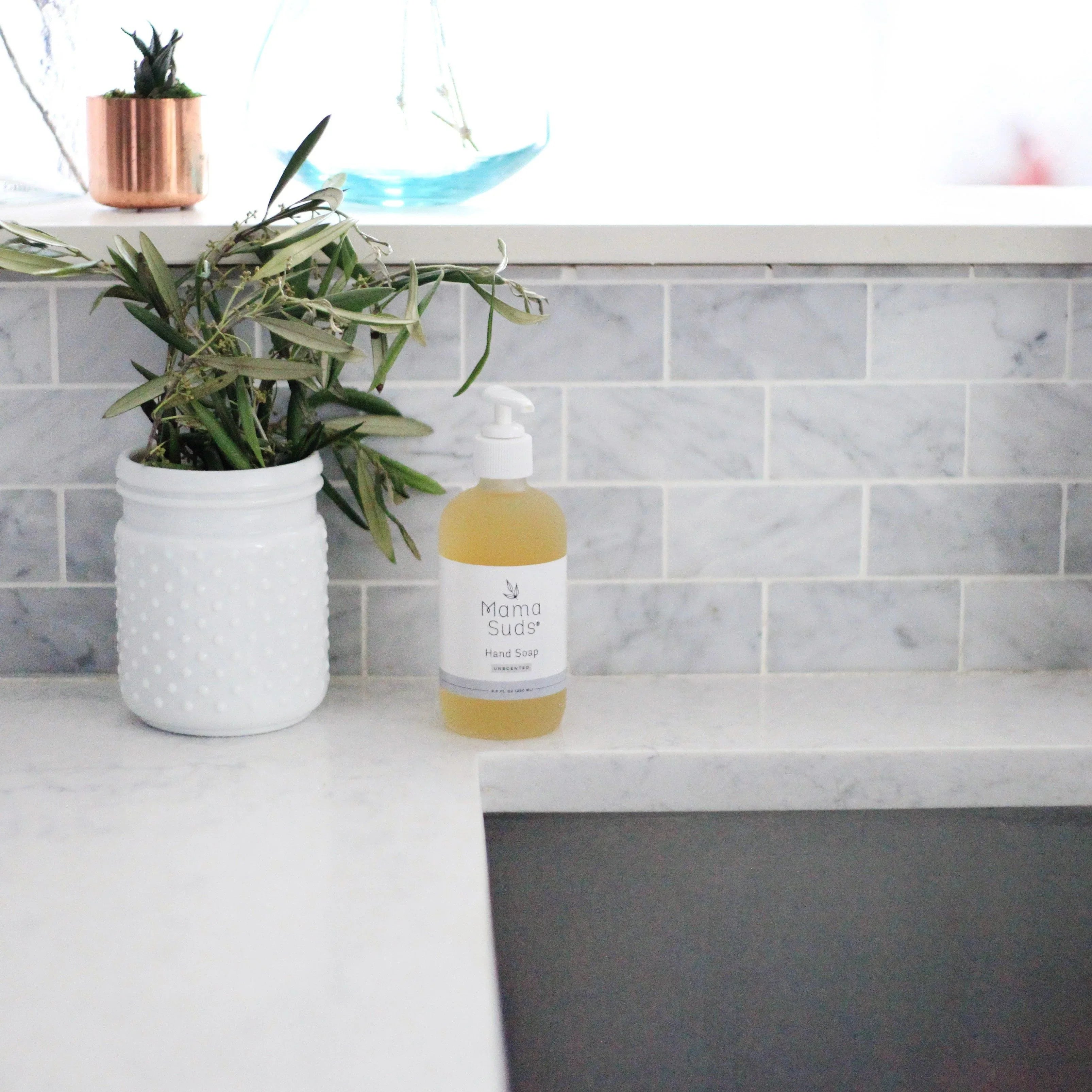 Mama Suds hand soap on white marble kitchen counter with plant, modern tile backsplash