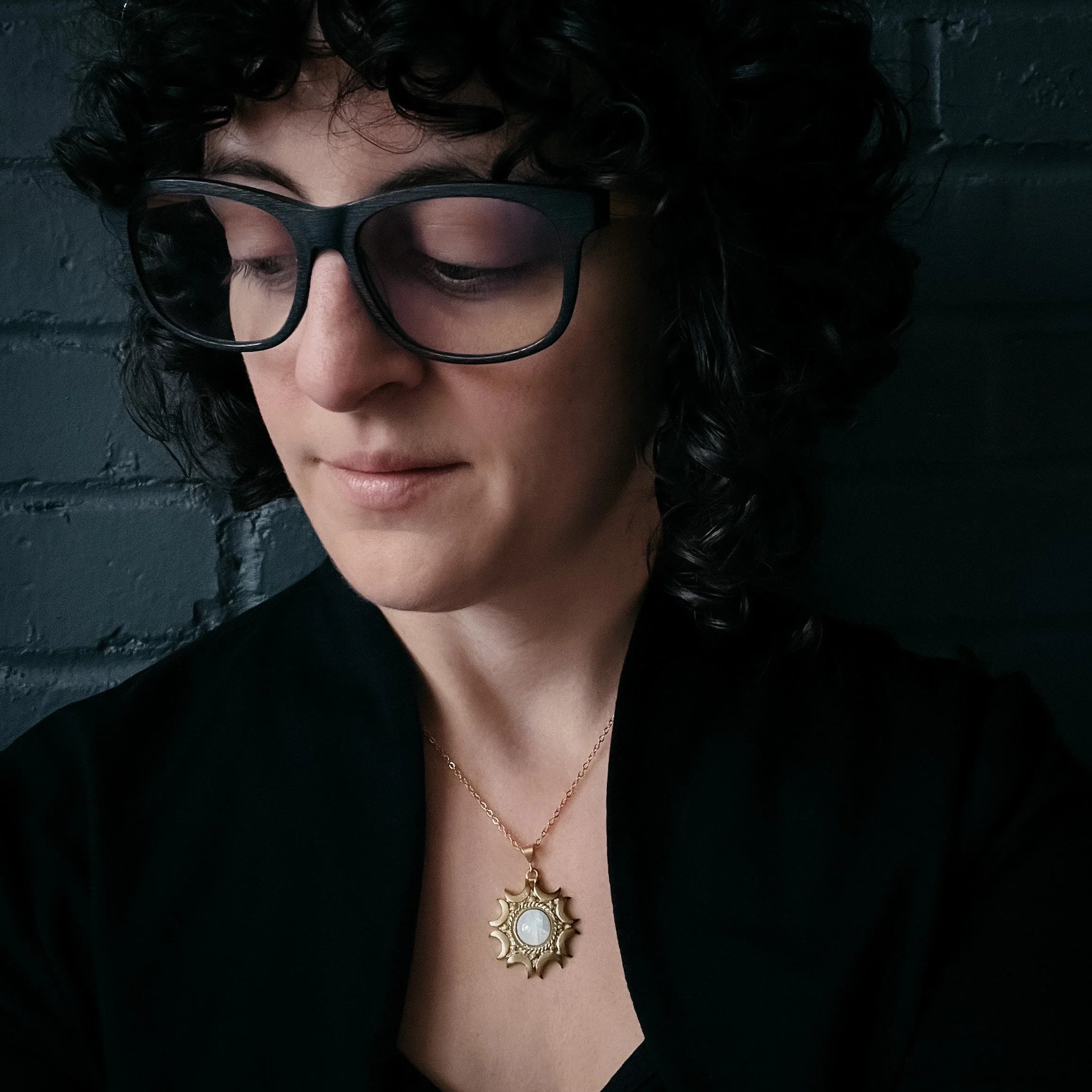 Woman with curly dark hair and black glasses wearing black clothes and a gold sun pendant necklace