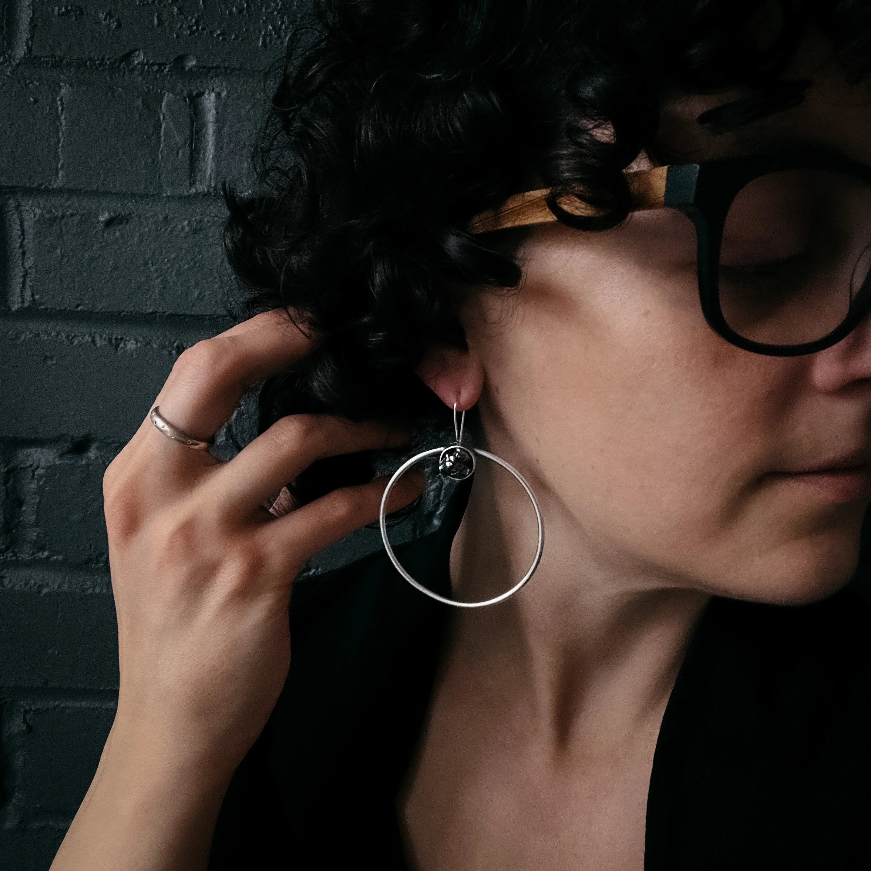 Close-up of woman with curly hair wearing large silver hoop earrings and black glasses against dark brick wall