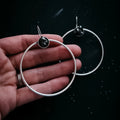 Hand holding a pair of silver hoop earrings with black stone accents on a dark starry background