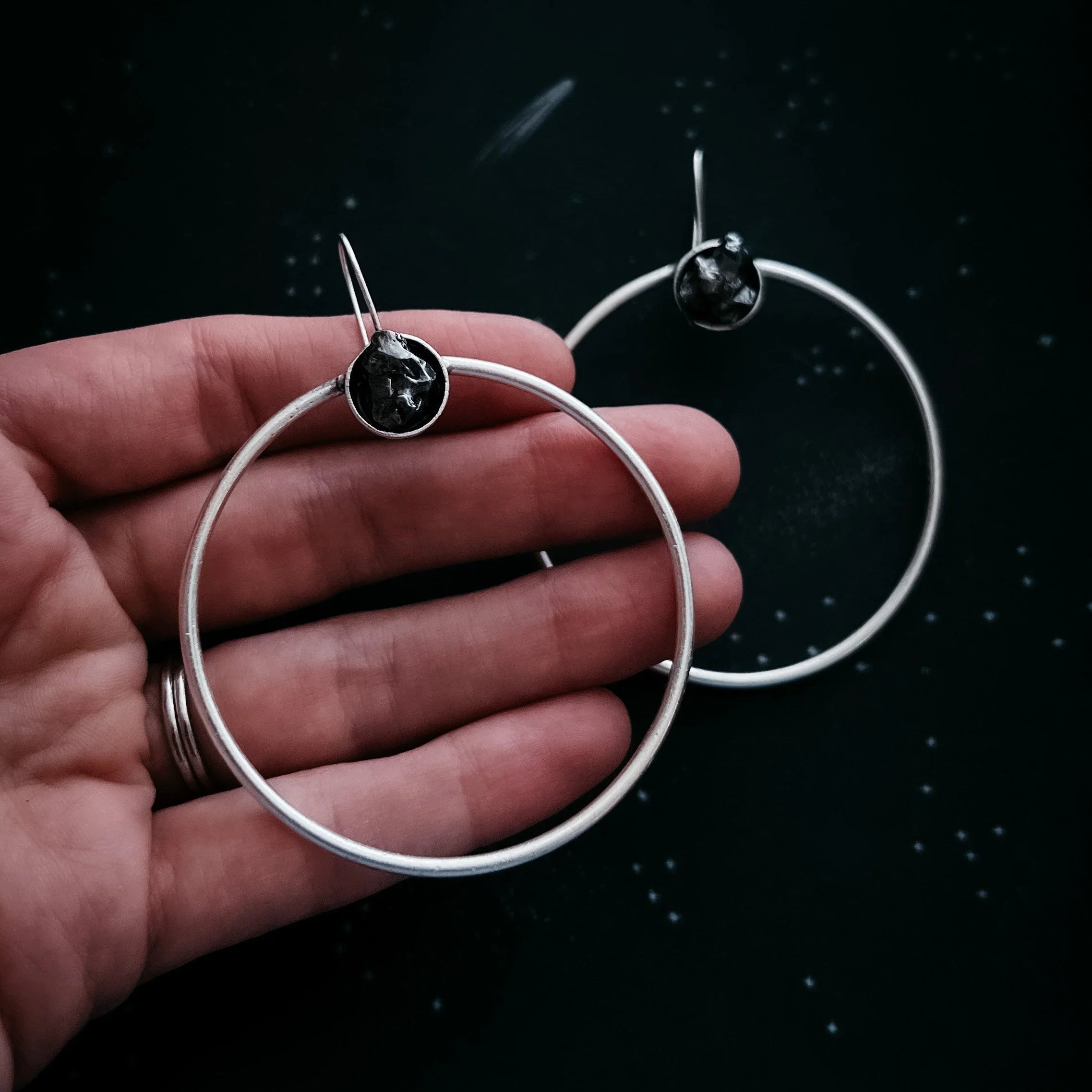 Hand holding a pair of silver hoop earrings with black stone accents on a dark starry background