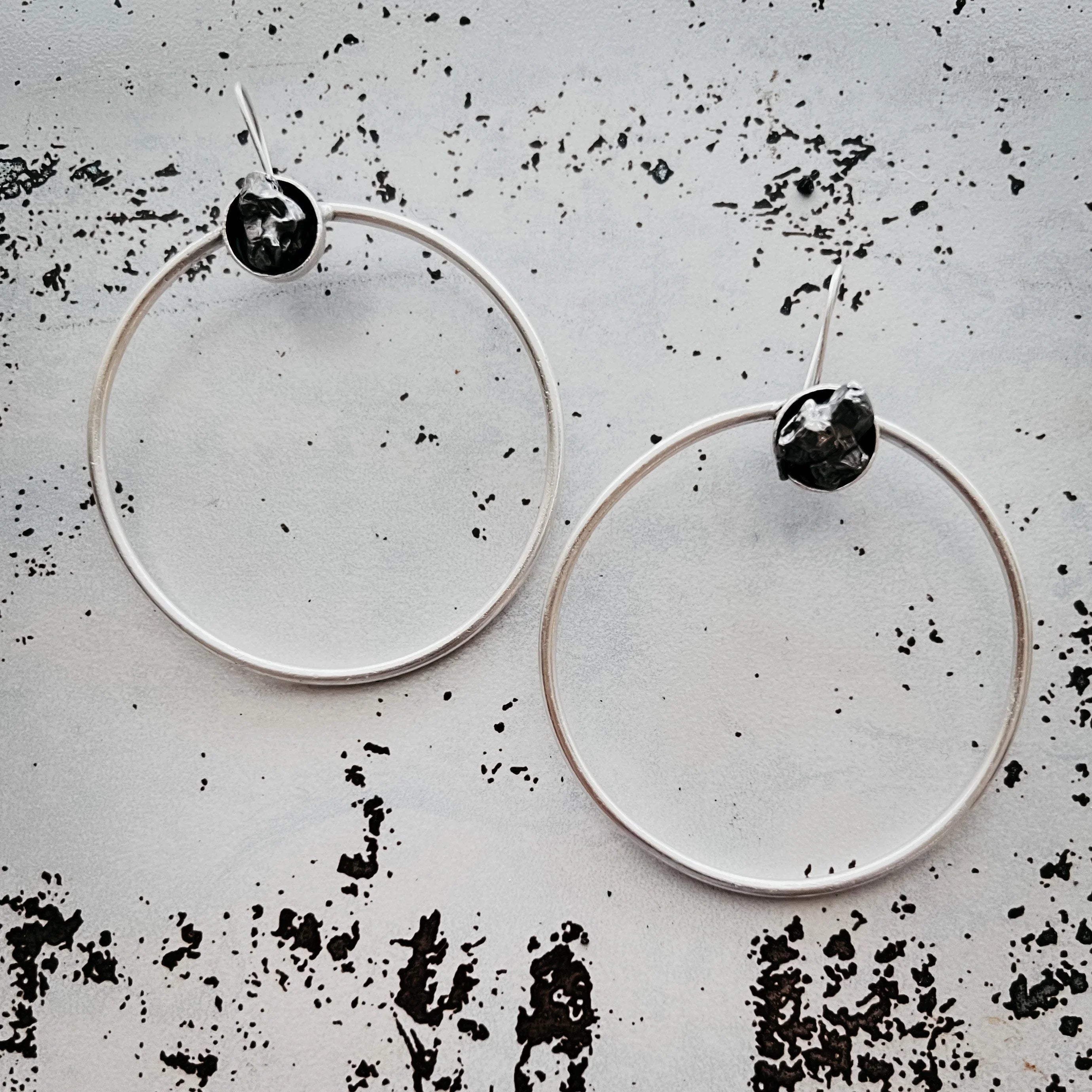 Minimalist silver hoop earrings with small hammered textured black accents on a distressed white background