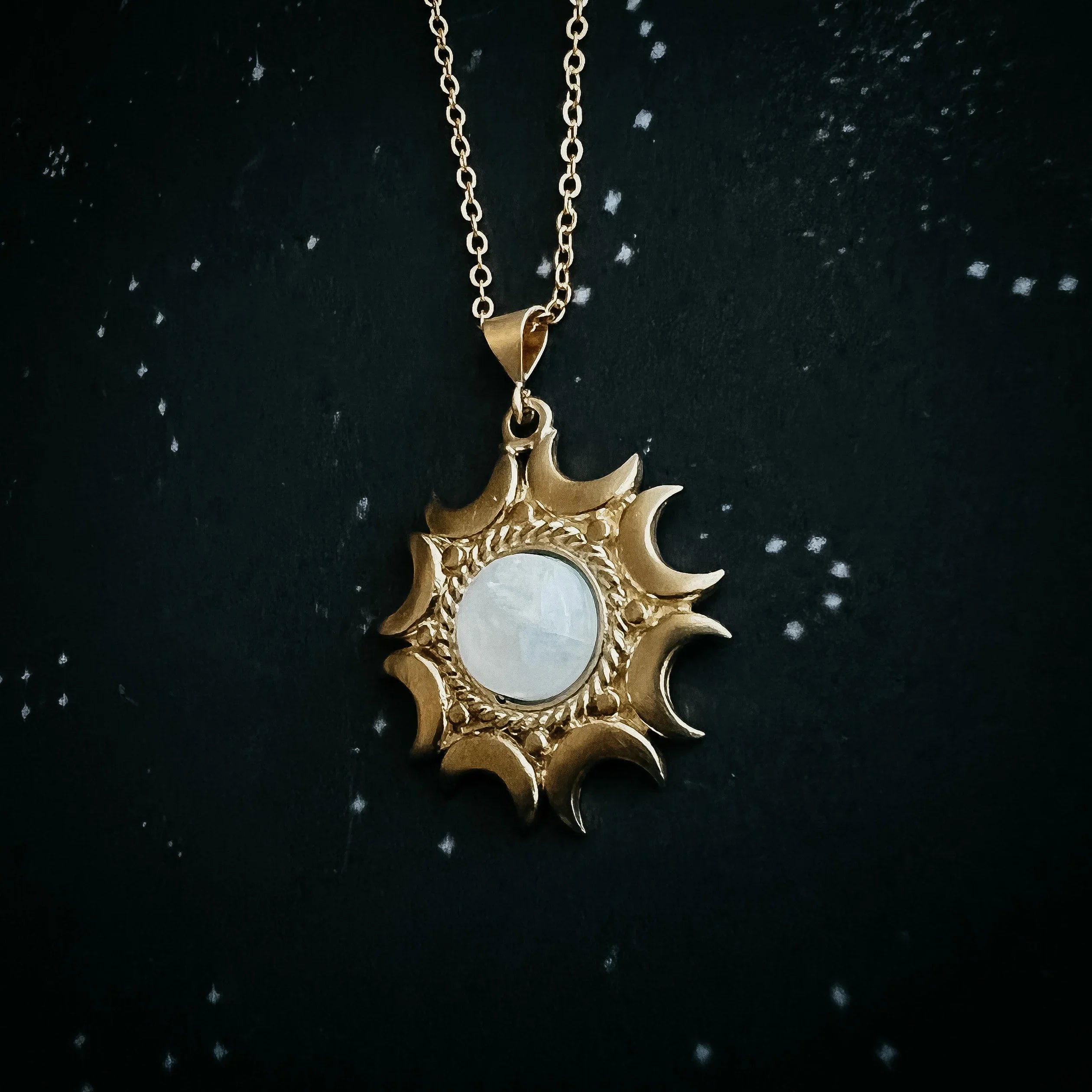 Gold necklace with sun and crescent moon pendant featuring a central white gemstone on black starry background