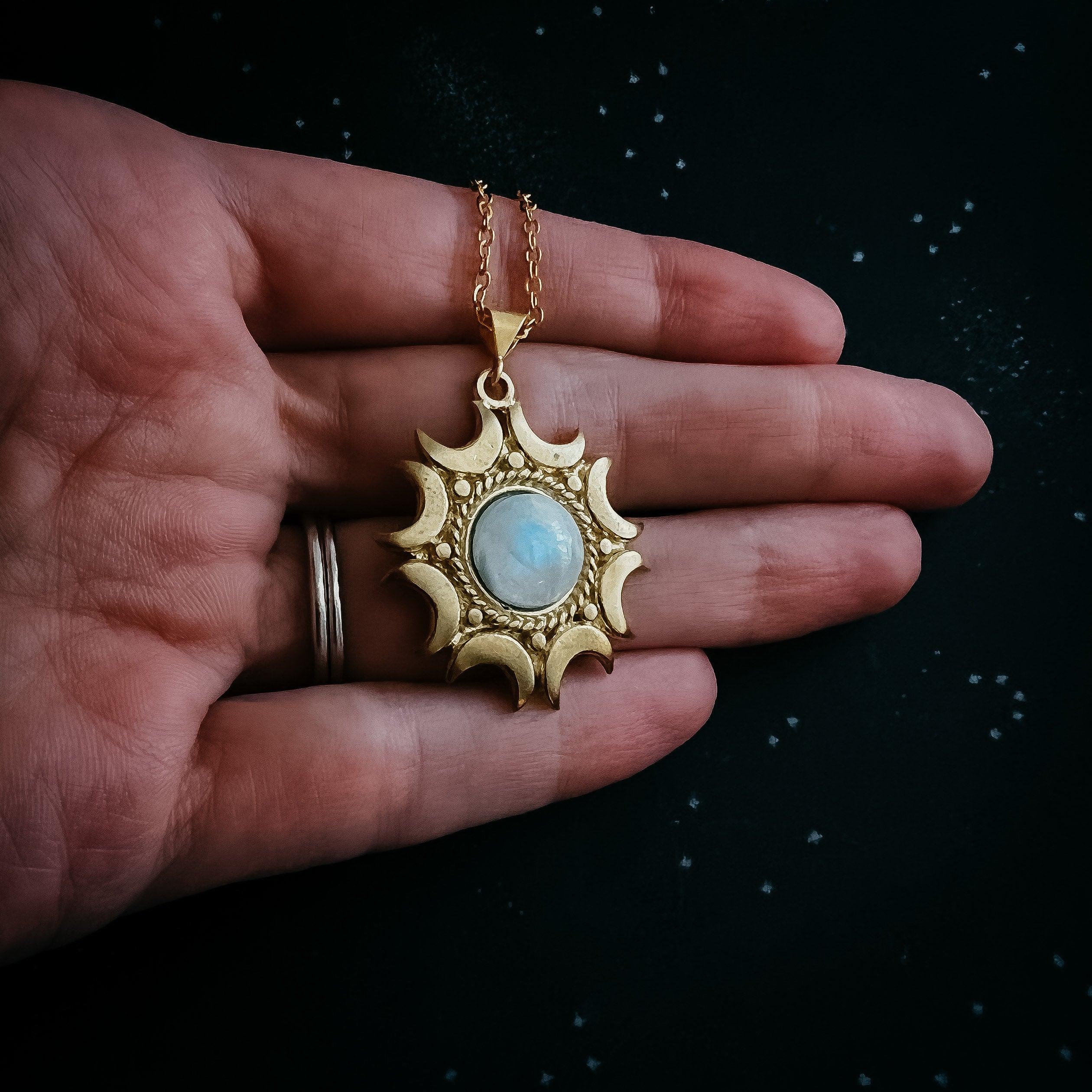 Hand holding gold sun pendant necklace with moon phases and blue gemstone center on dark background