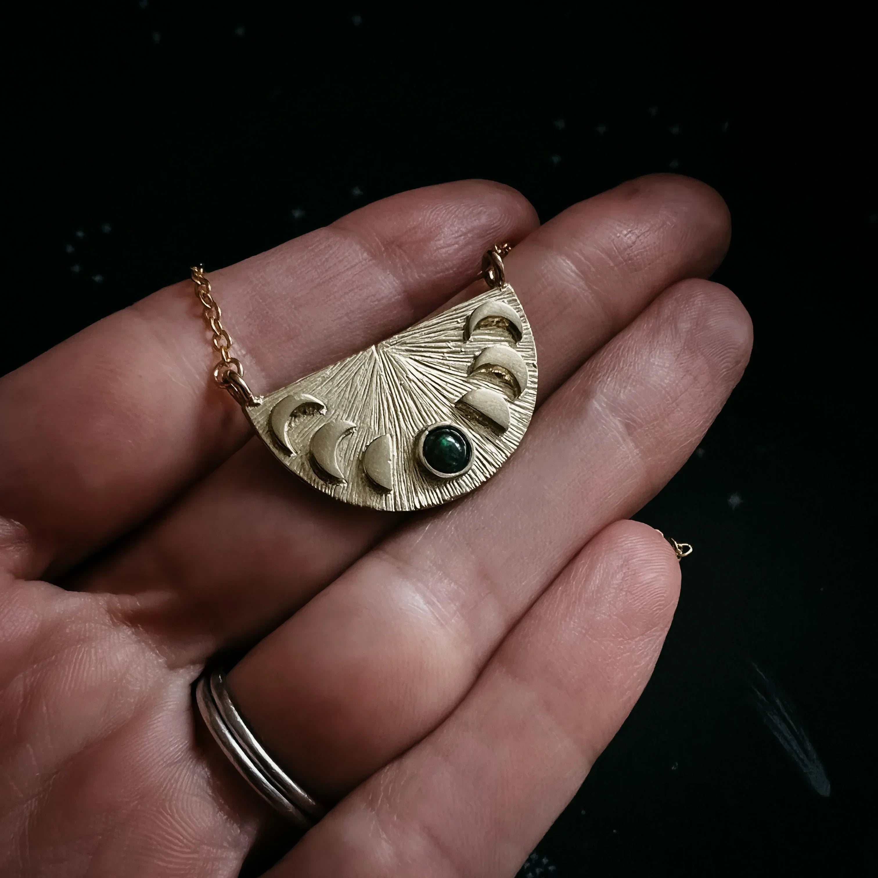 Hand holding gold moon phase necklace pendant with green stone accent on black background