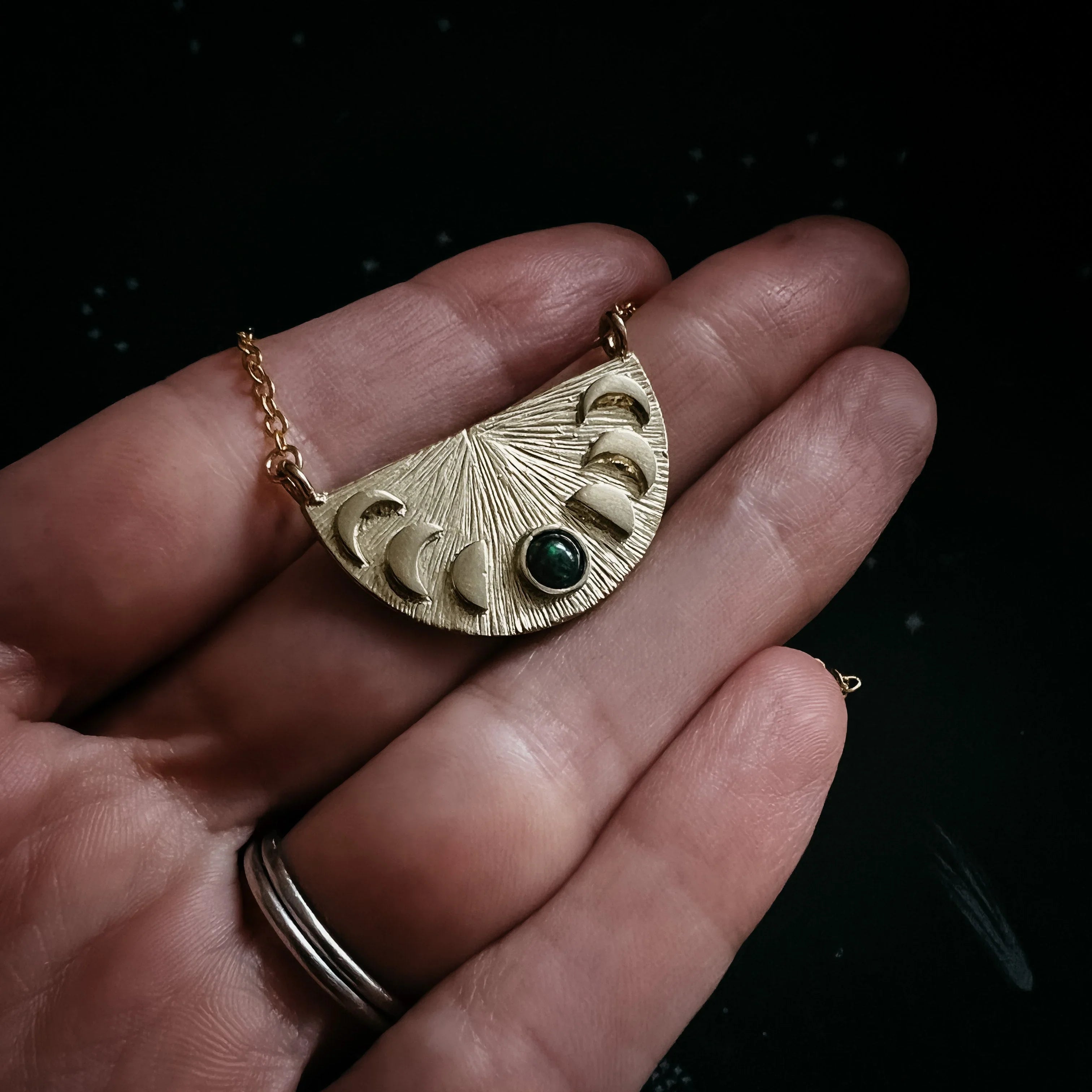 Hand holding gold crescent moon phase necklace pendant with green gemstone on dark background