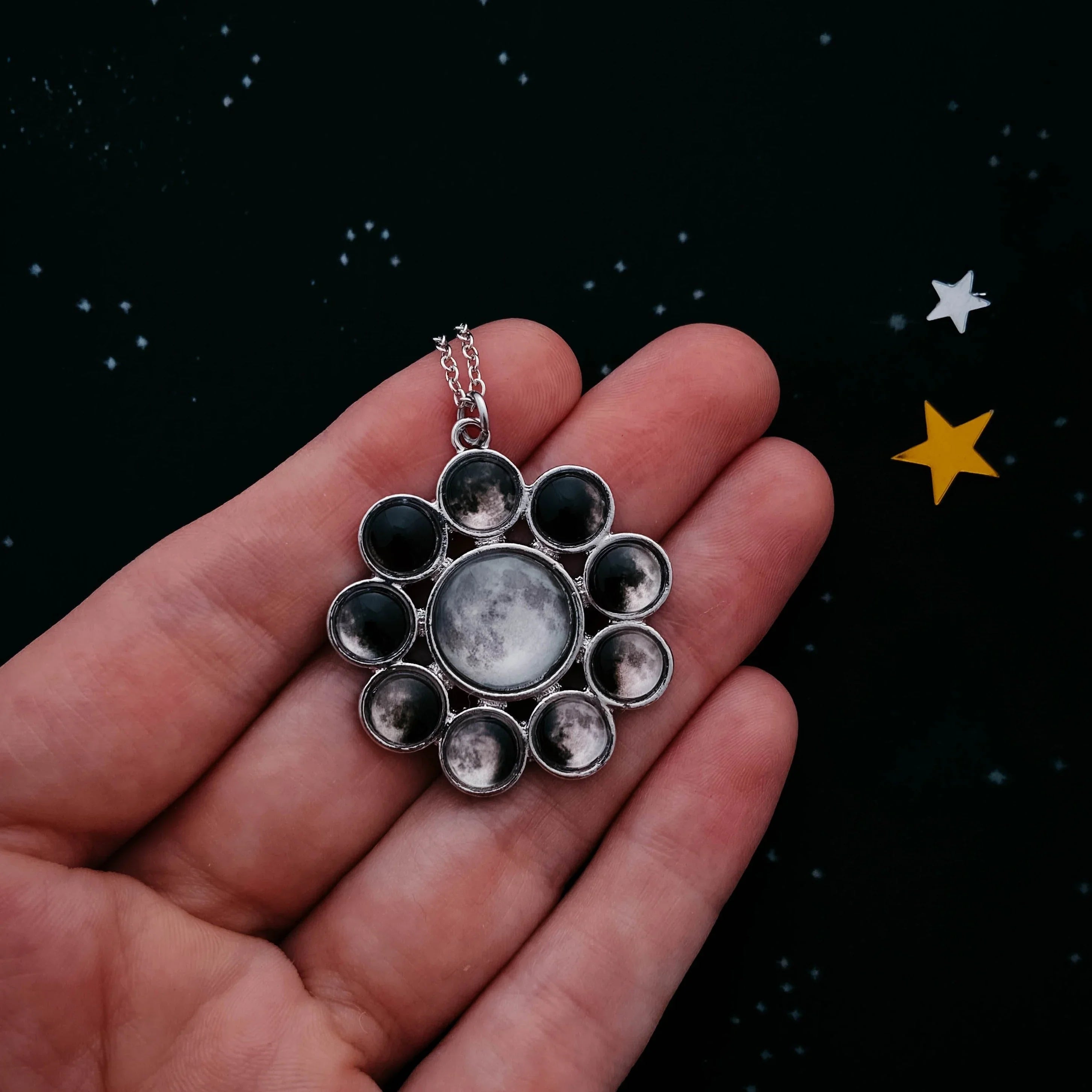 Hand holding a silver moon phase pendant necklace against a starry black background