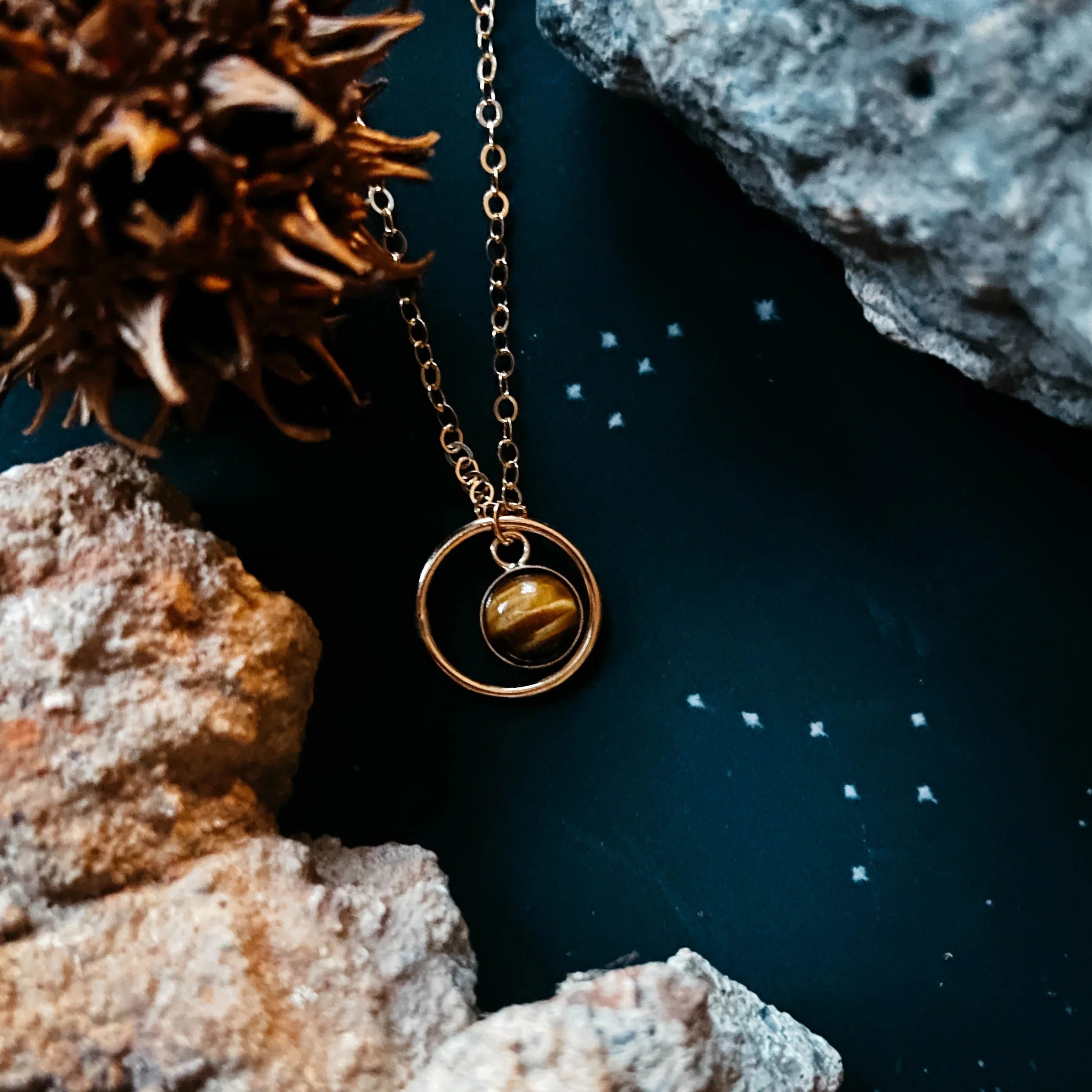 Gold pendant necklace with tiger's eye stone on dark surface with stones, cosmic boutique style