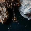Gold pendant necklace with brown stone, styled on black surface with rocks and dried seed pod, Qu!rk Boutique