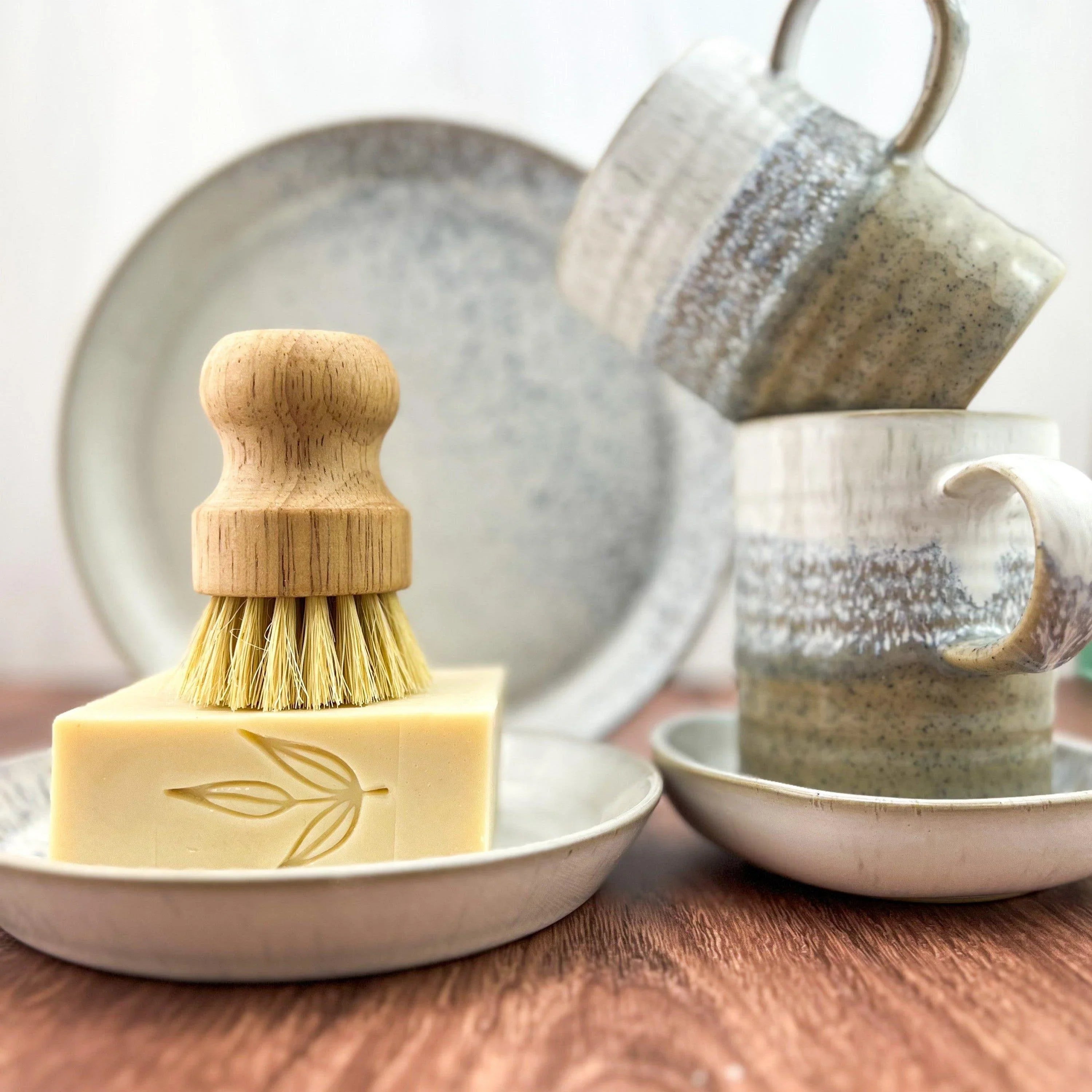 Handcrafted ceramic mugs, eco-friendly dish brush on artisan soap, and stoneware plates on wood table at Qu!rk Boutique.