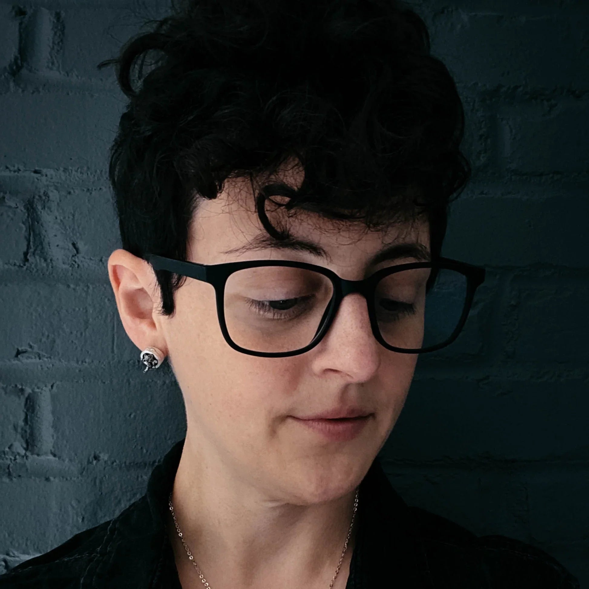 Person with short curly hair wearing black glasses and silver earrings against dark brick wall
