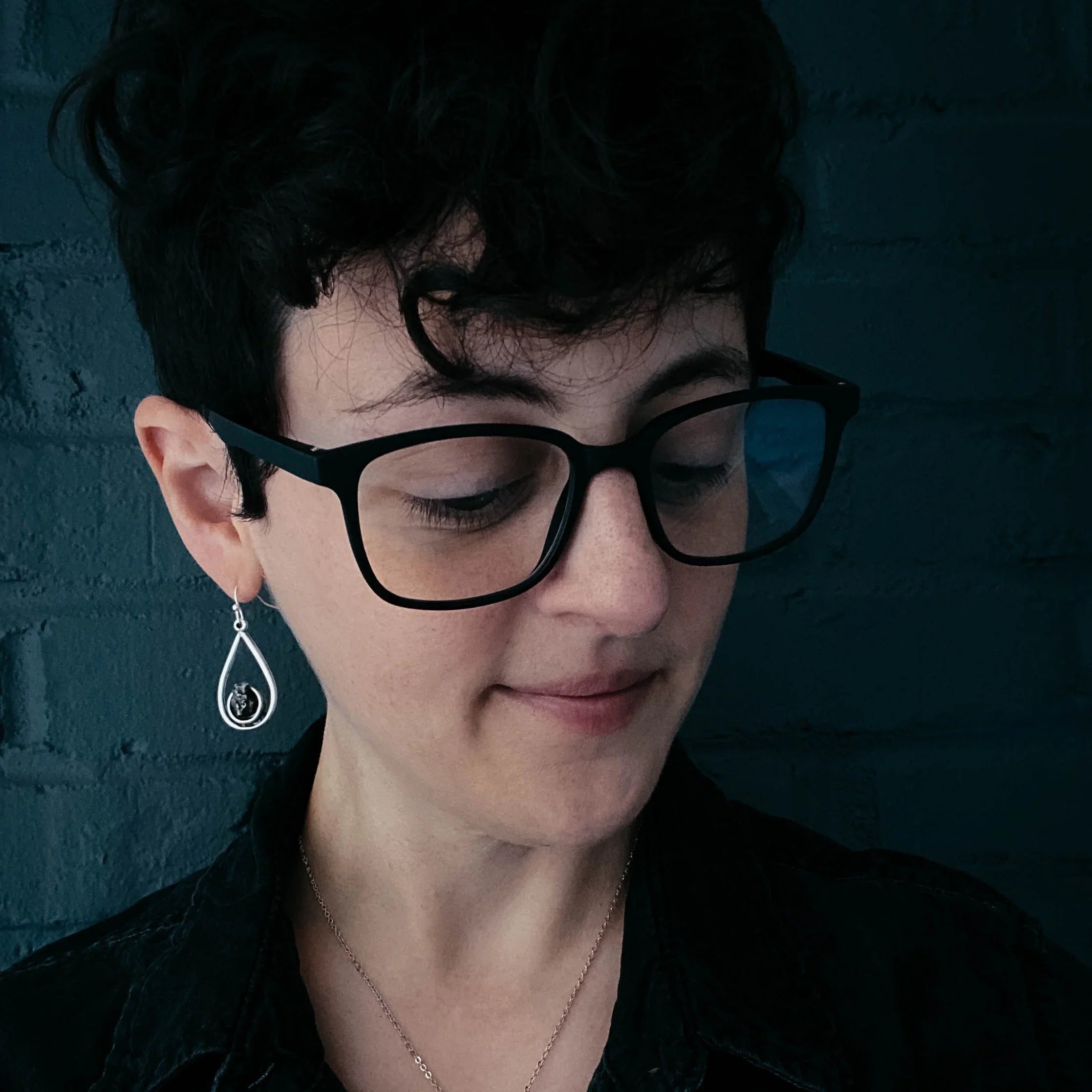 Person with short curly hair wearing black glasses, teardrop earring, and dark clothing against dark brick wall