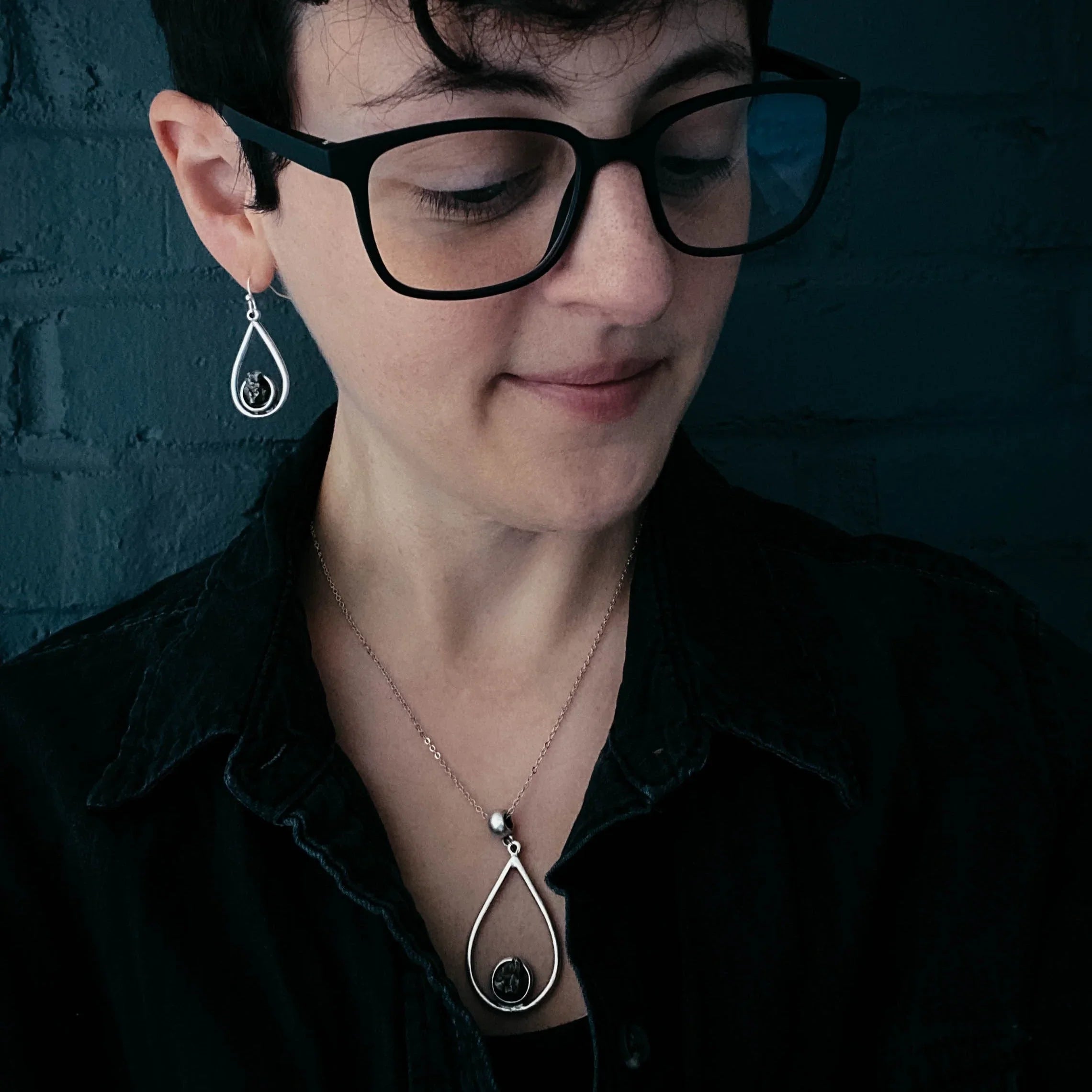 Person with short hair and black glasses wearing matching silver teardrop earrings and necklace against dark background