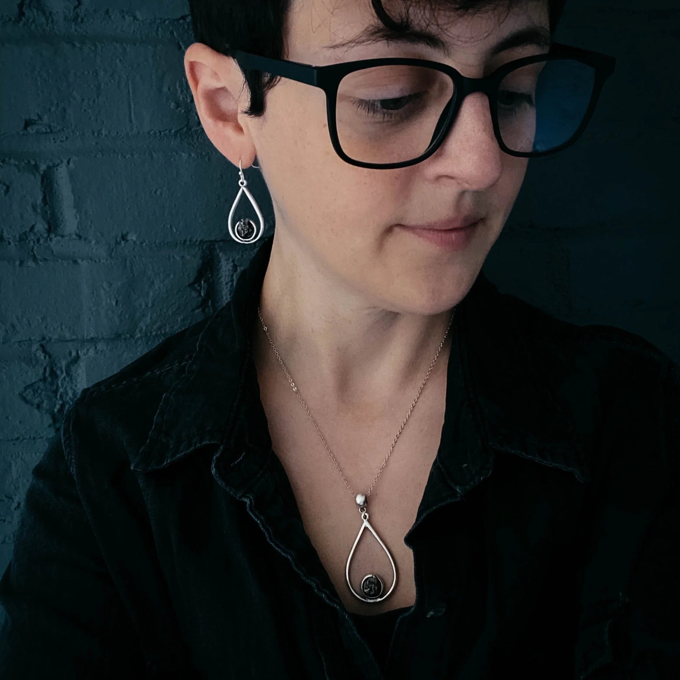 Woman wearing matching silver teardrop earrings and pendant necklace with black details, black shirt, and glasses