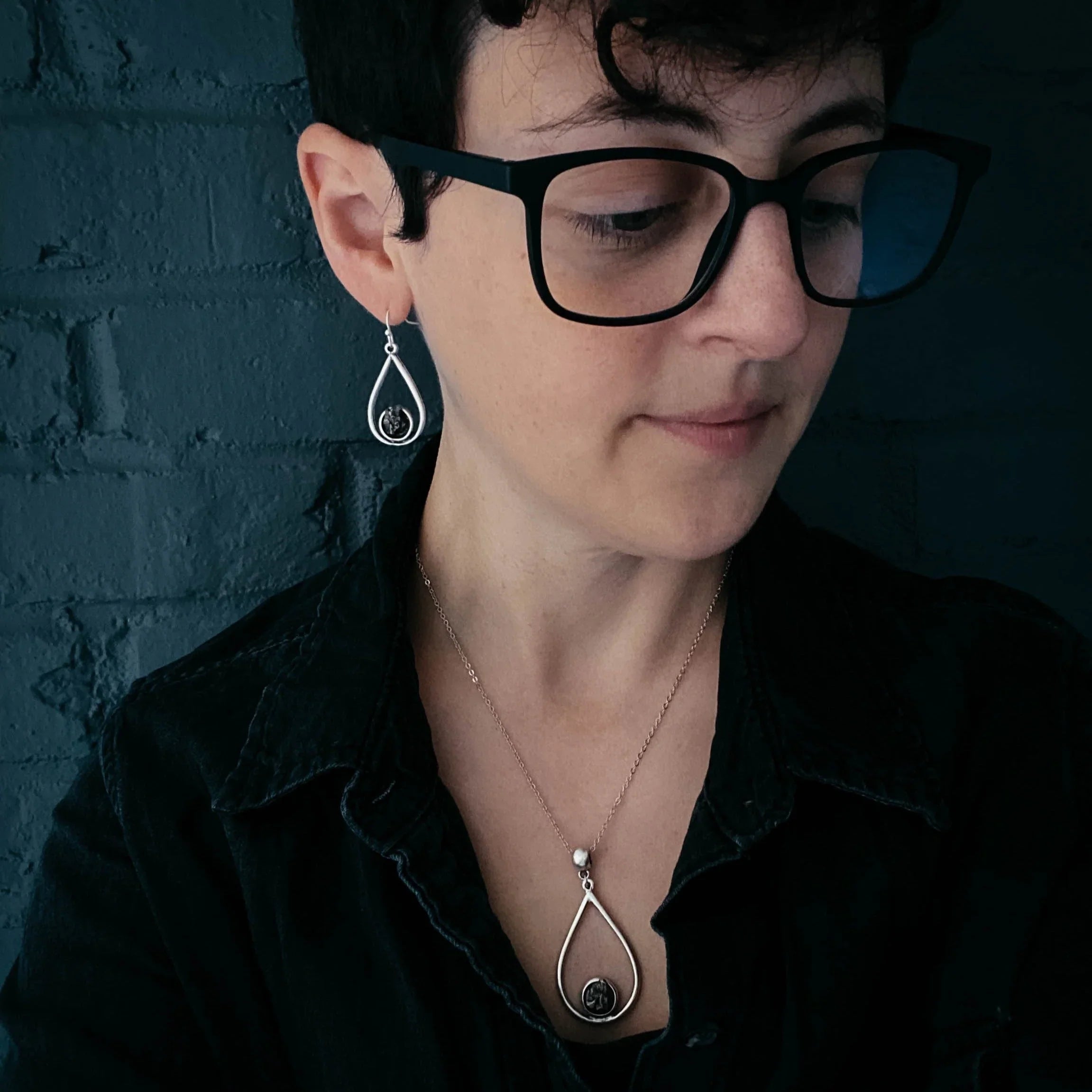 Person wearing black eyeglasses and black shirt with matching silver teardrop earrings and pendant necklace