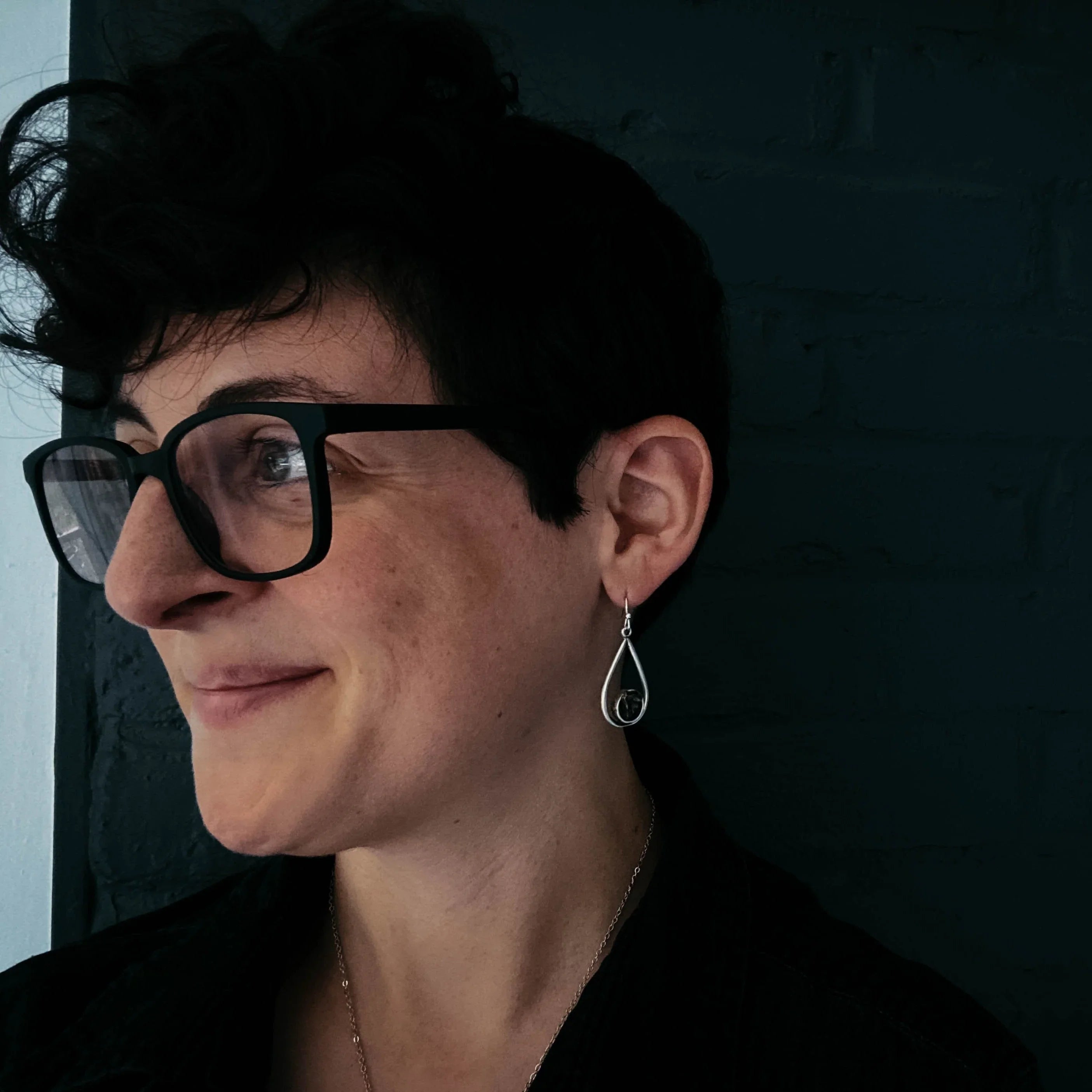Close-up of person with short curly hair wearing black glasses and teardrop silver earrings against dark brick wall