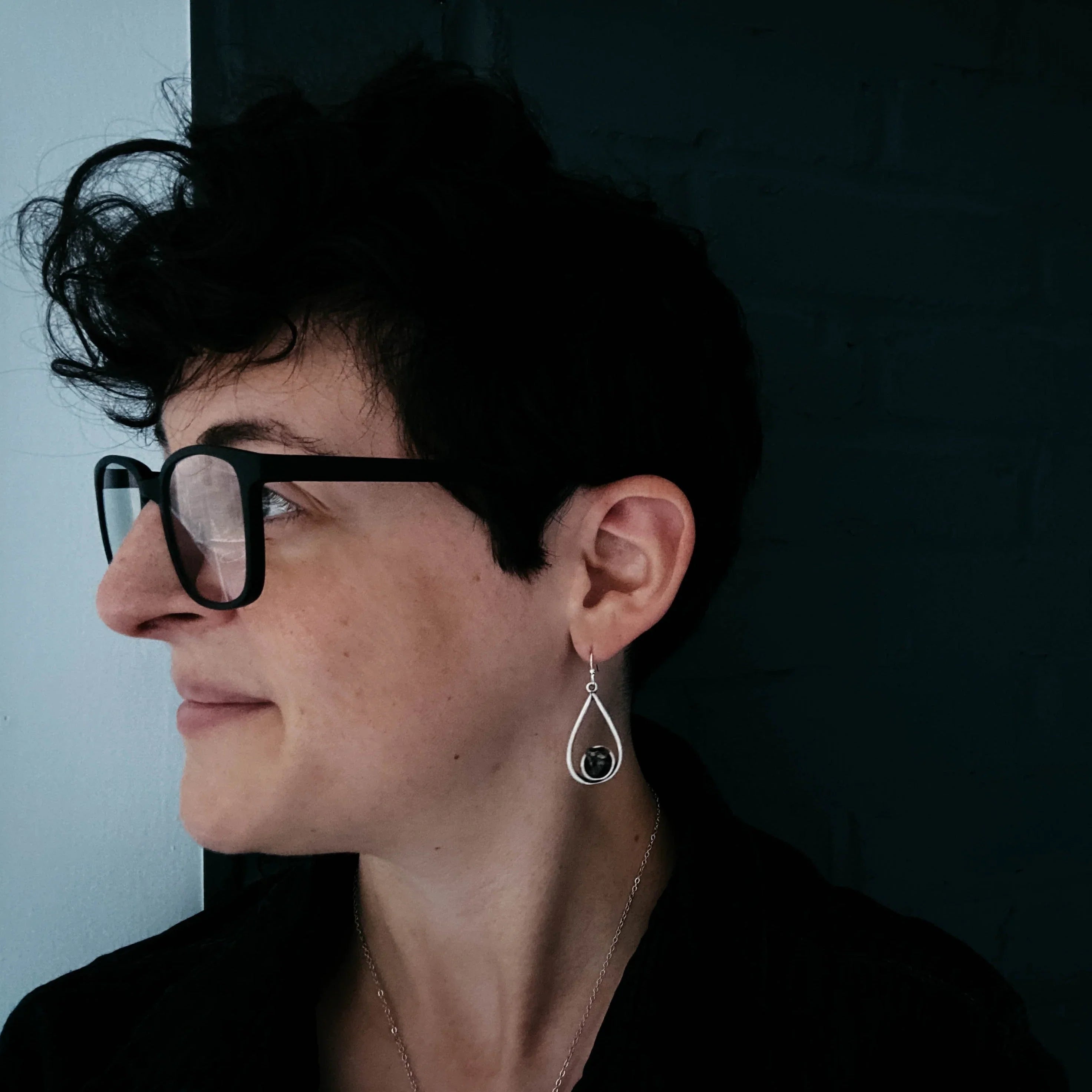 Person with short curly hair and black rectangular glasses wearing silver teardrop earrings with black stone